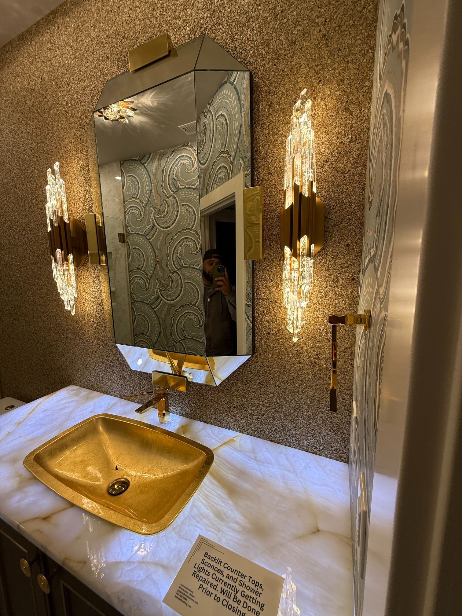 Gold sink and wall mirror in a tiled bathroom with a marble countertop and wall sconces.