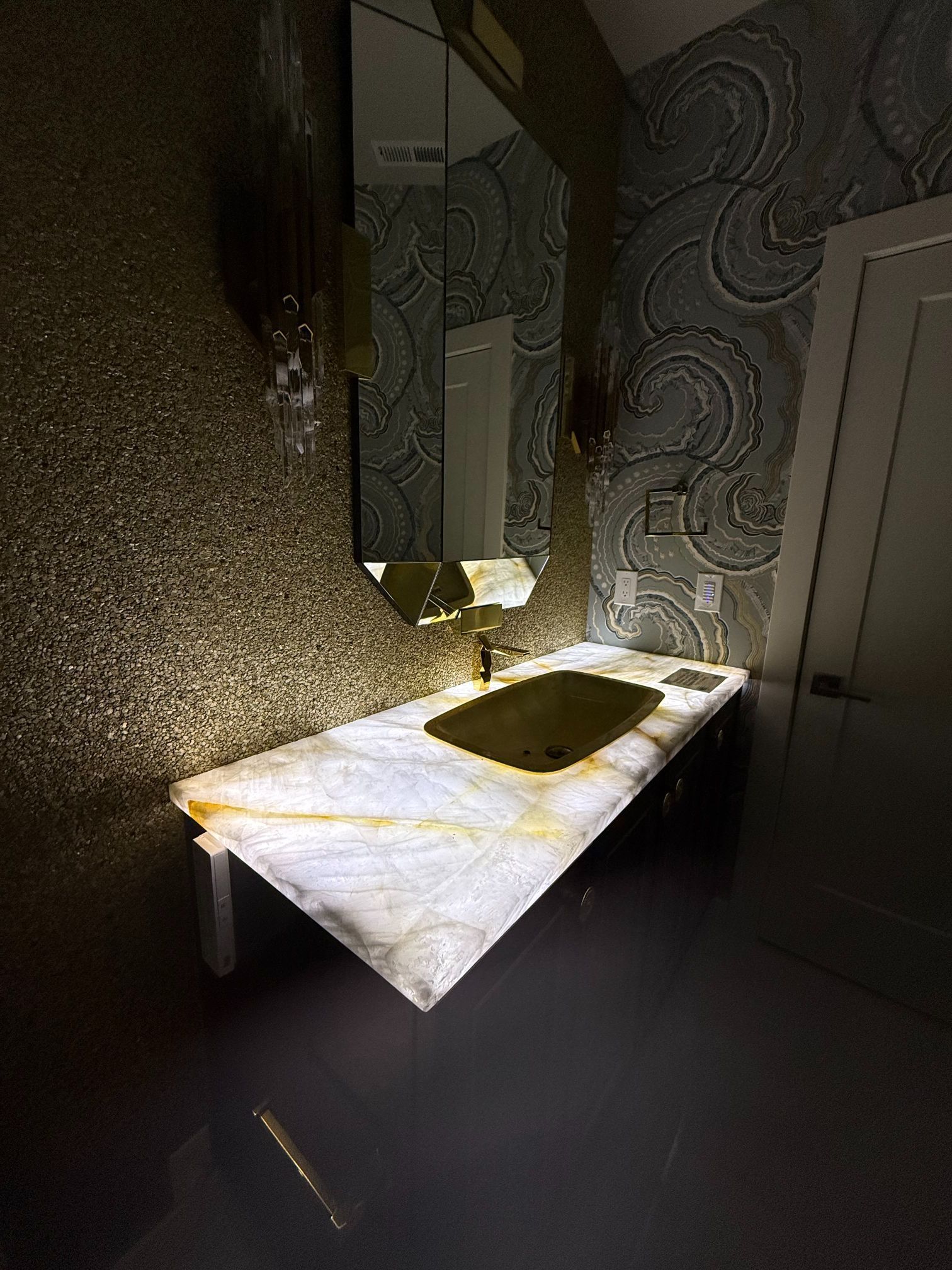 Dim bathroom with a marble sink, wall mirror, and patterned wallpaper in shadow.