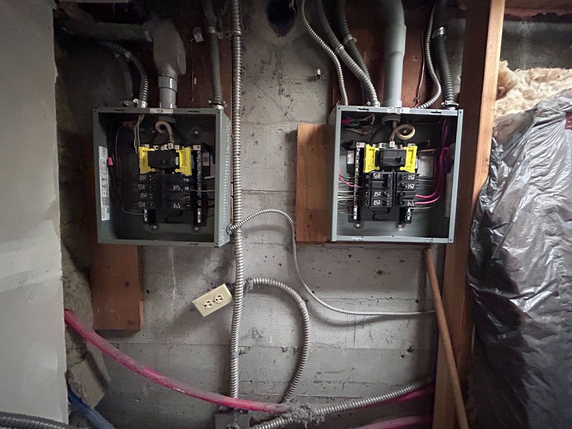 Two open electrical breaker panels on a concrete wall with visible wiring and pipes in a utility area