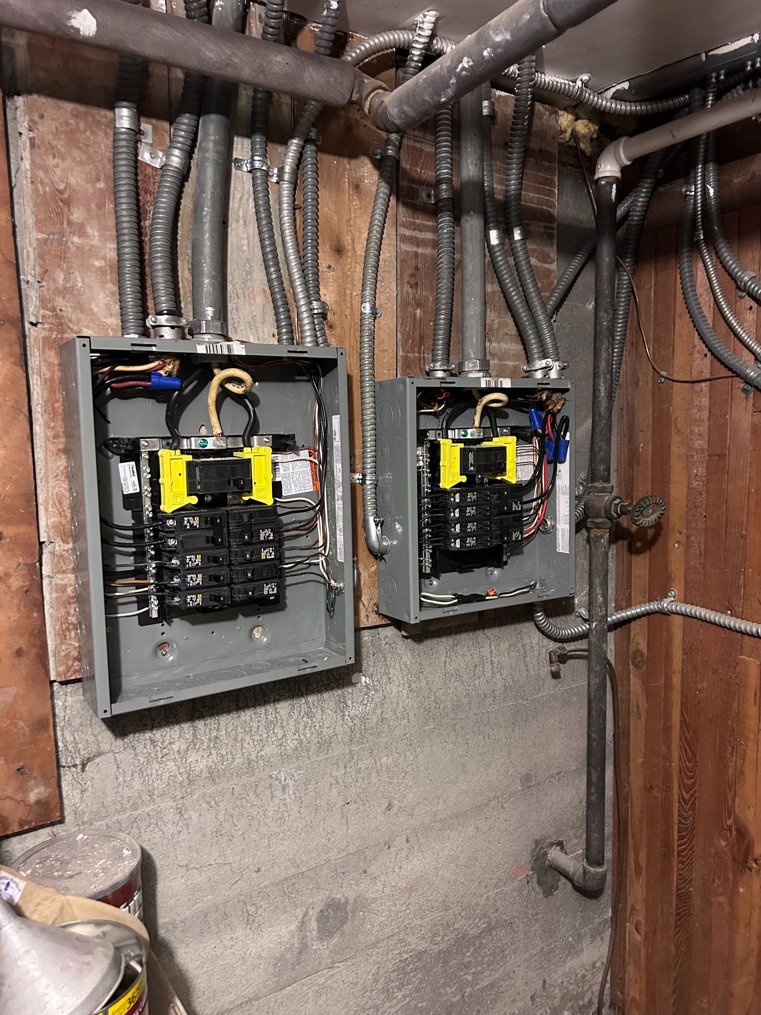 Two open electrical breaker panels with exposed wiring in an unfinished basement wall