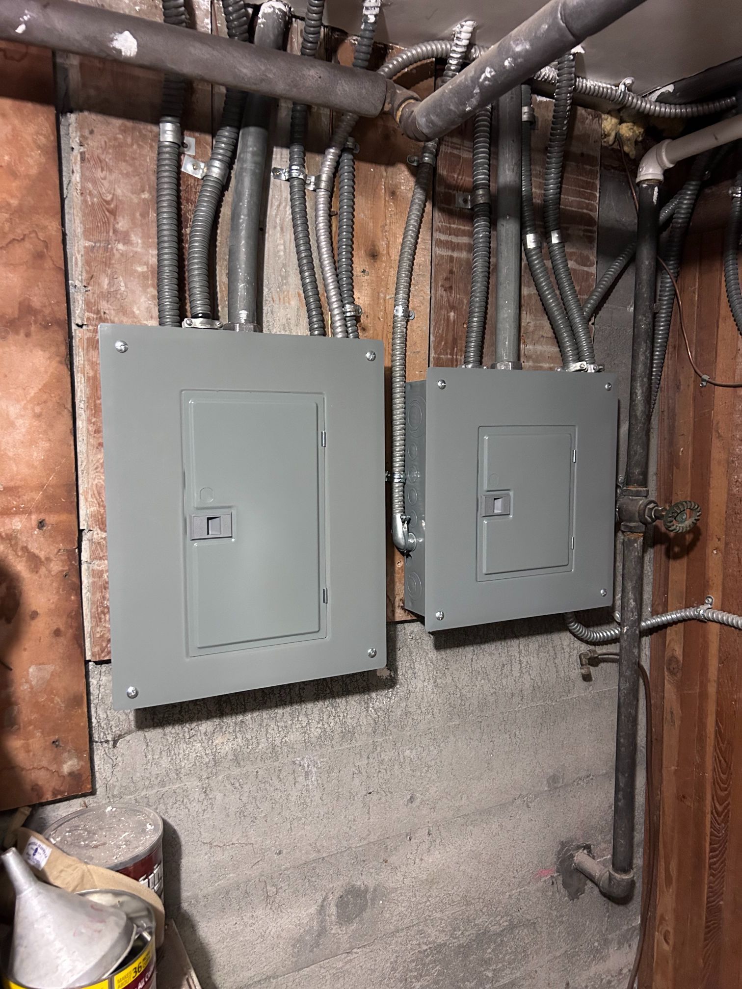 Two gray electrical panels mounted on a basement wall with exposed wiring above them
