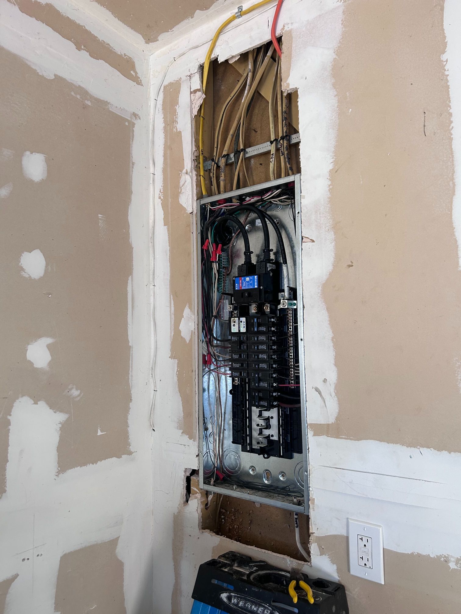 Exposed electrical panel in a drywall wall during home renovation, with visible wiring and breakers.