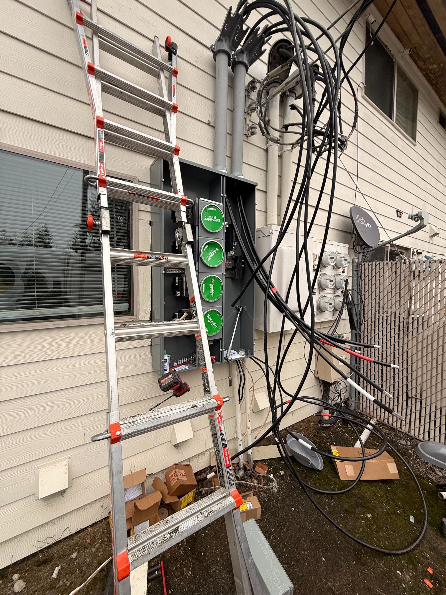 Aluminum ladder beside a house exterior, with cables, wall-mounted equipment, and a window nearby.