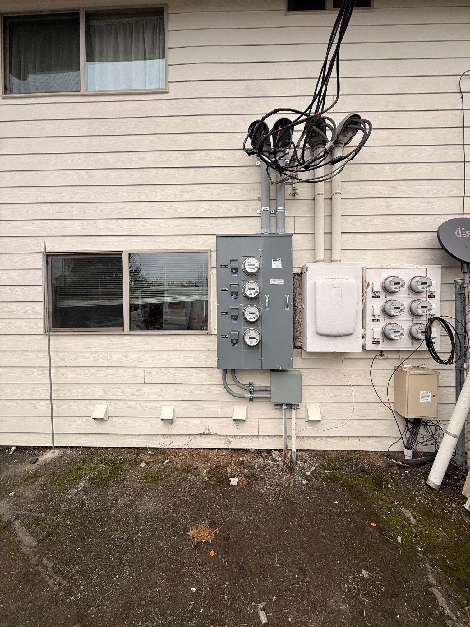 Exterior wall with utility meters, electrical panels, and overhead cables beside a gravel yard.
