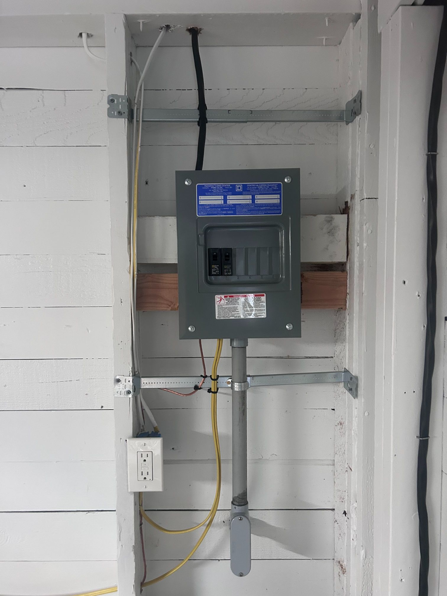 Electrical breaker panel in a gray utility closet with exposed wiring and conduit attached to the wall.