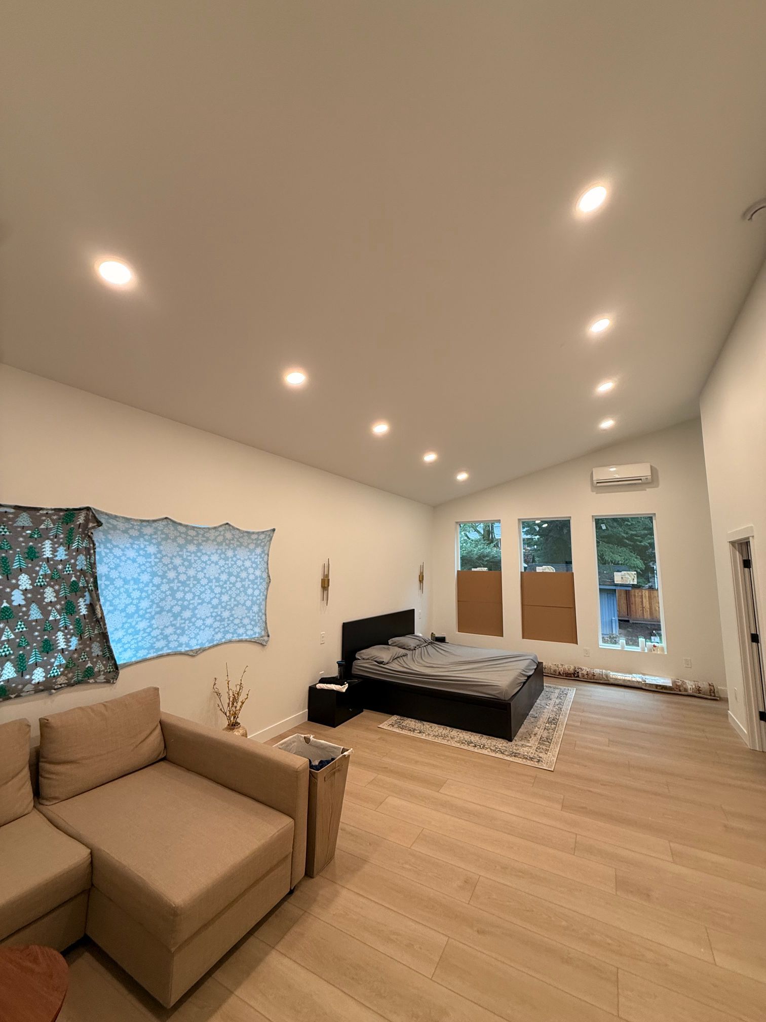 Spacious beige bedroom with a sofa, bed, windows with floral curtains, and recessed ceiling lights.