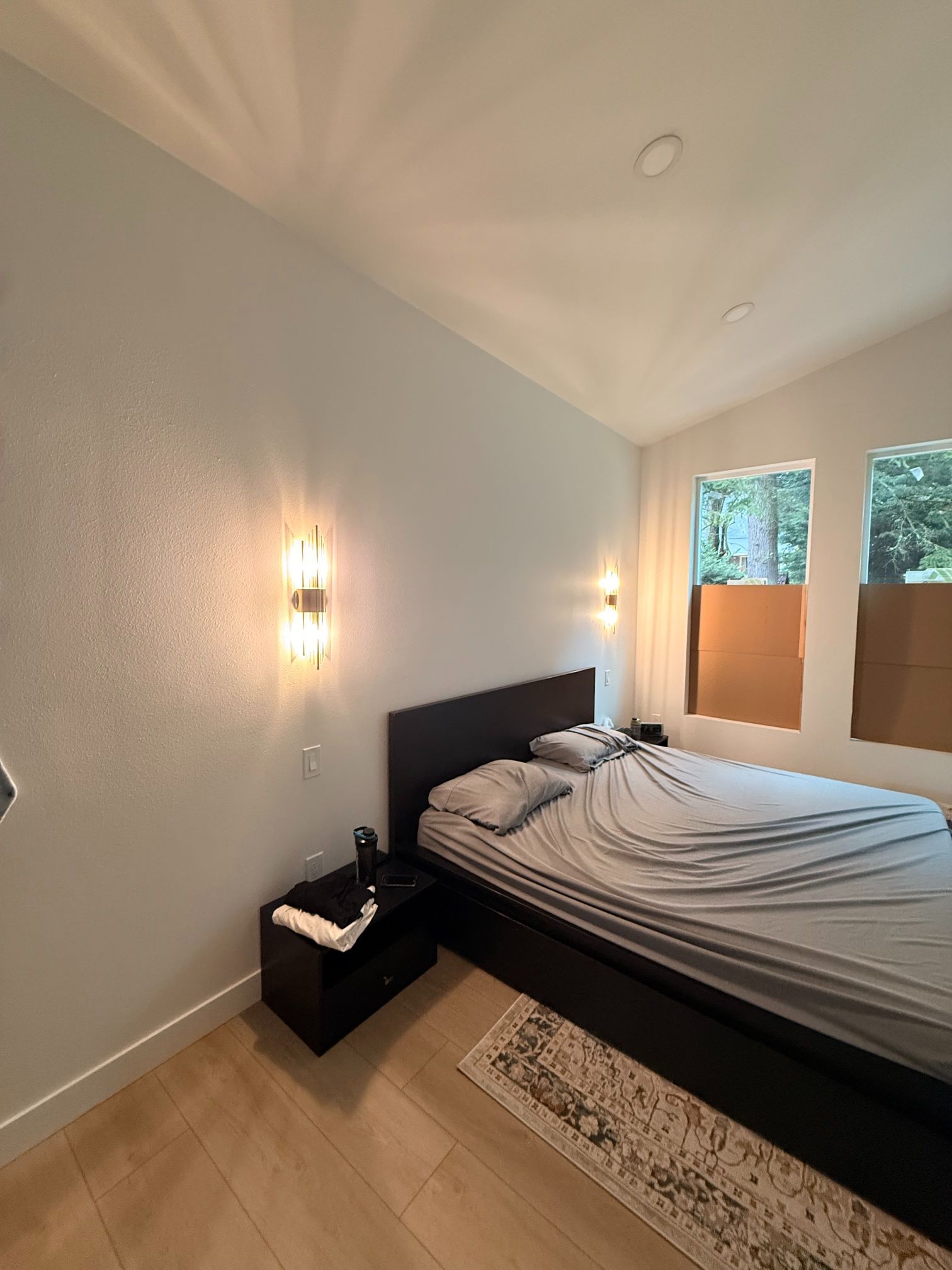 Minimalist bedroom with unmade bed, wall sconces, and two windows with blinds