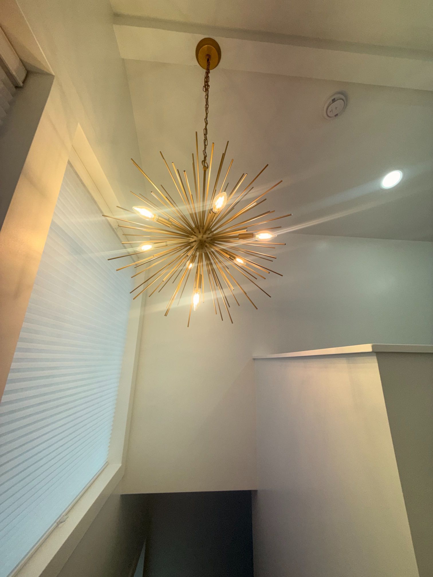 Gold starburst chandelier hanging in a white stairwell with a skylight and window blinds