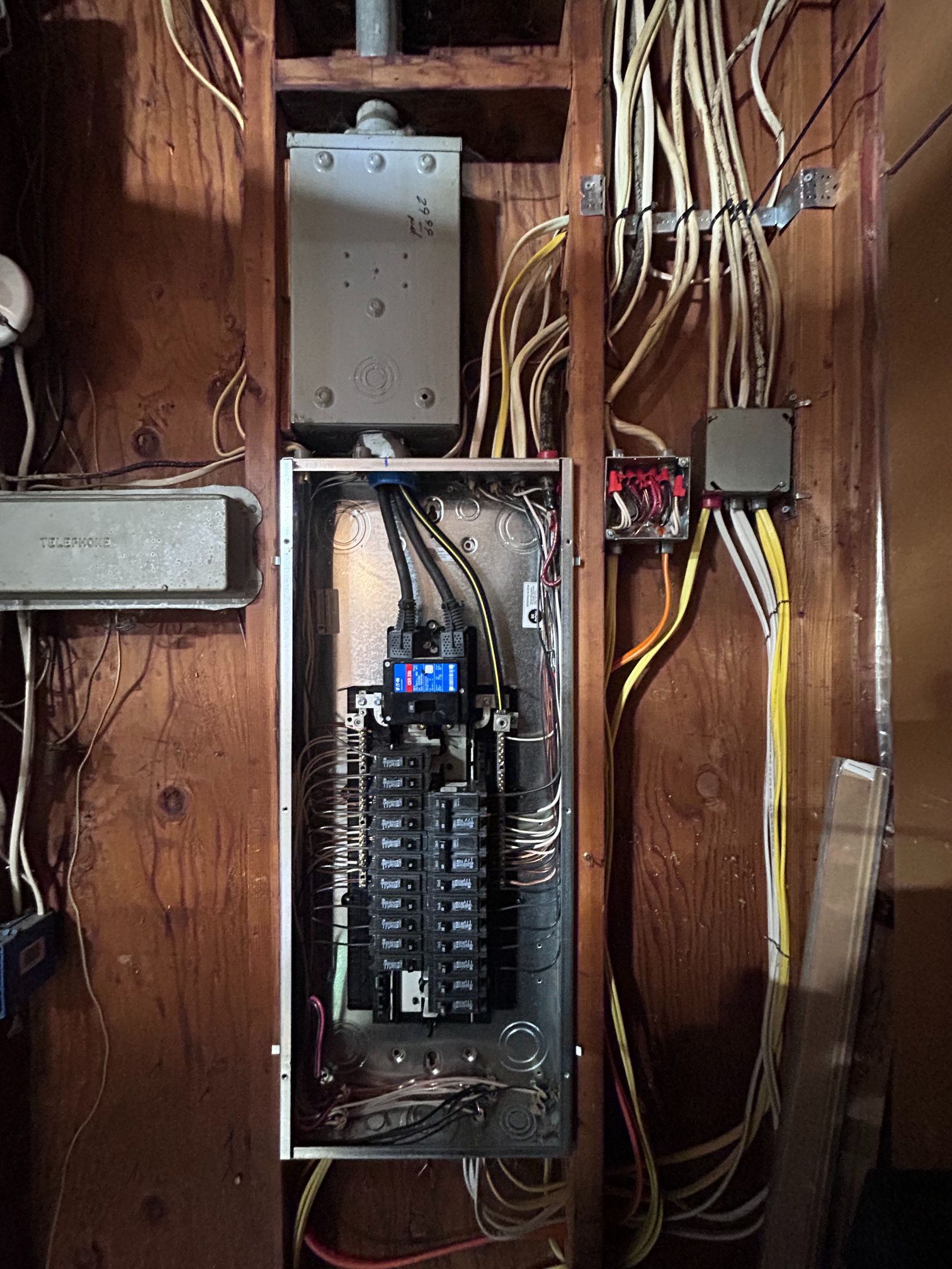 Open electrical panel with breakers and exposed wiring in a wooden wall cavity
