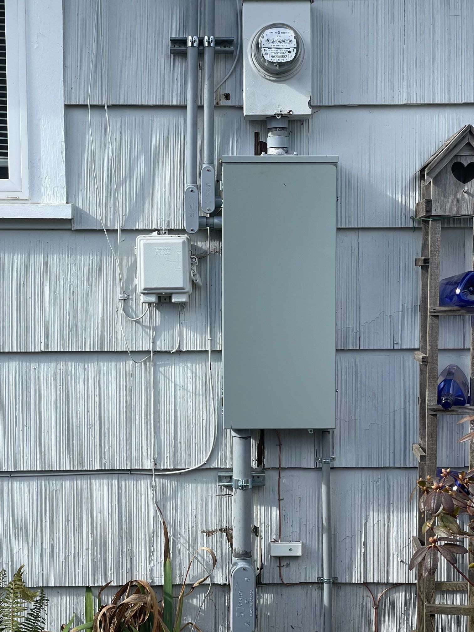 Gray electrical meter and breaker box mounted on the exterior wall of a house.
