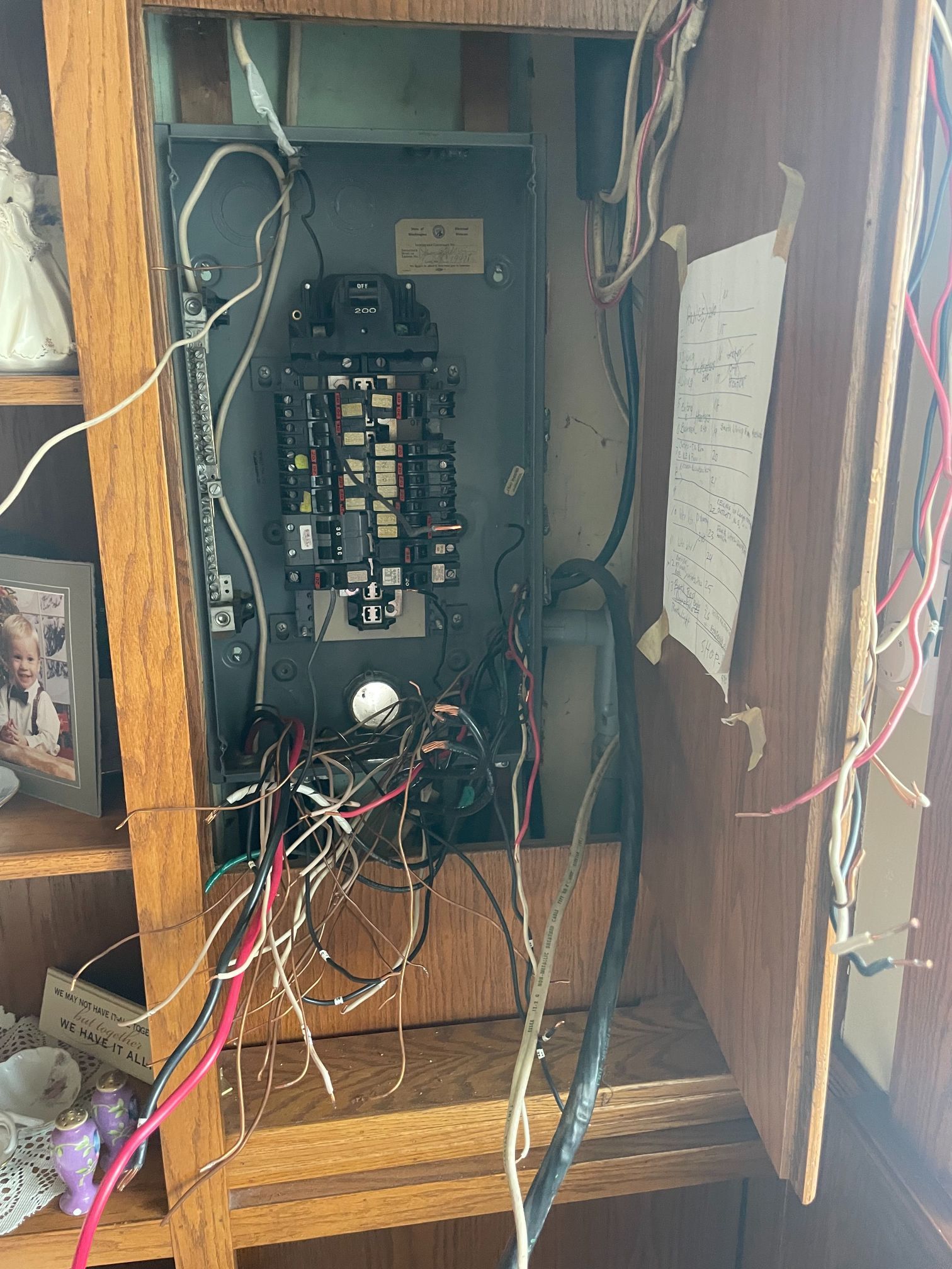 Open electrical breaker panel with exposed wires inside a wooden wall cabinet.