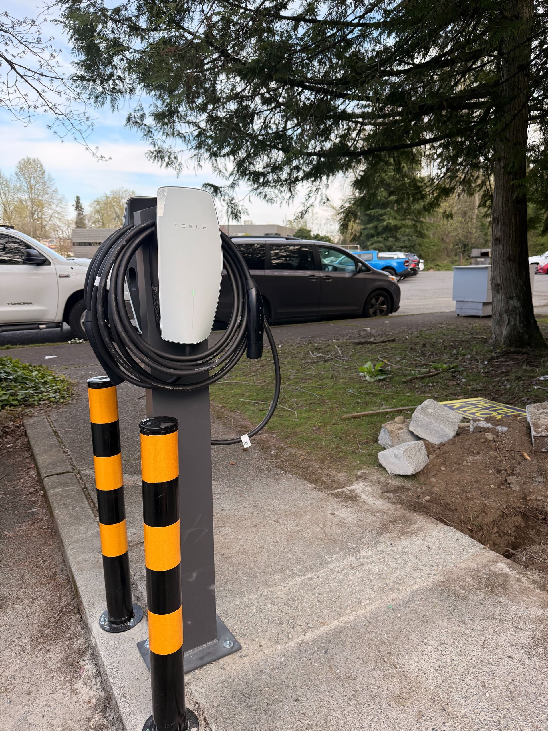 Outdoor EV charging station with coiled cable on a post beside parked cars and trees