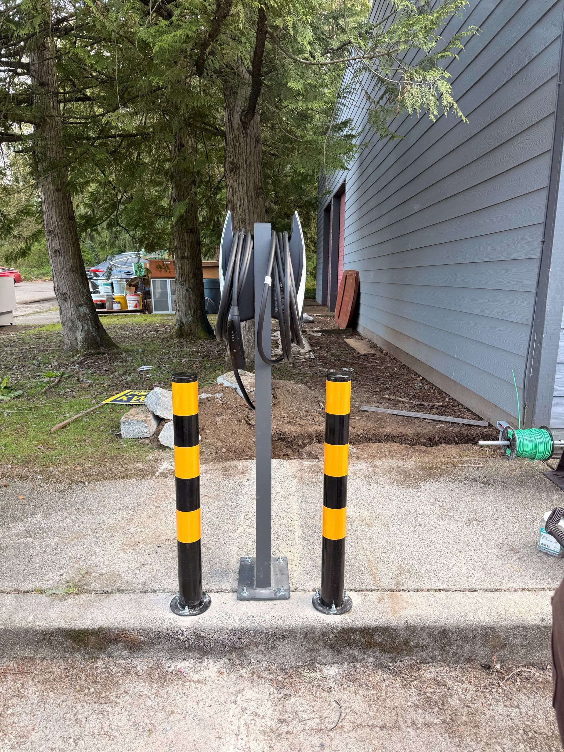 Black-and-yellow bollards guard a metal bike rack beside a building and trees on a gravel path.