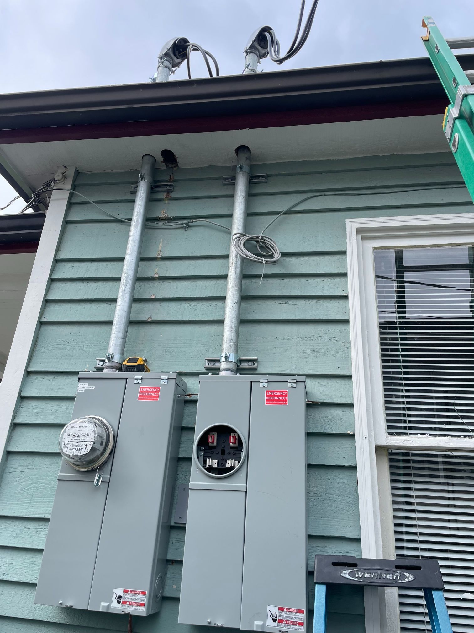 Gray electrical meter boxes mounted on light green siding under a roof eave, with a window on the right