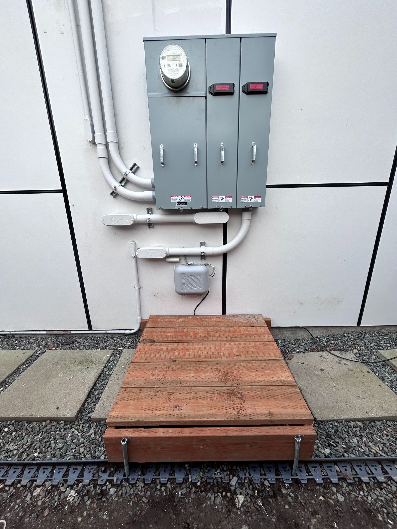 Outdoor utility meters and gray electrical boxes above a wooden platform walkway, with gravel and paving stones below