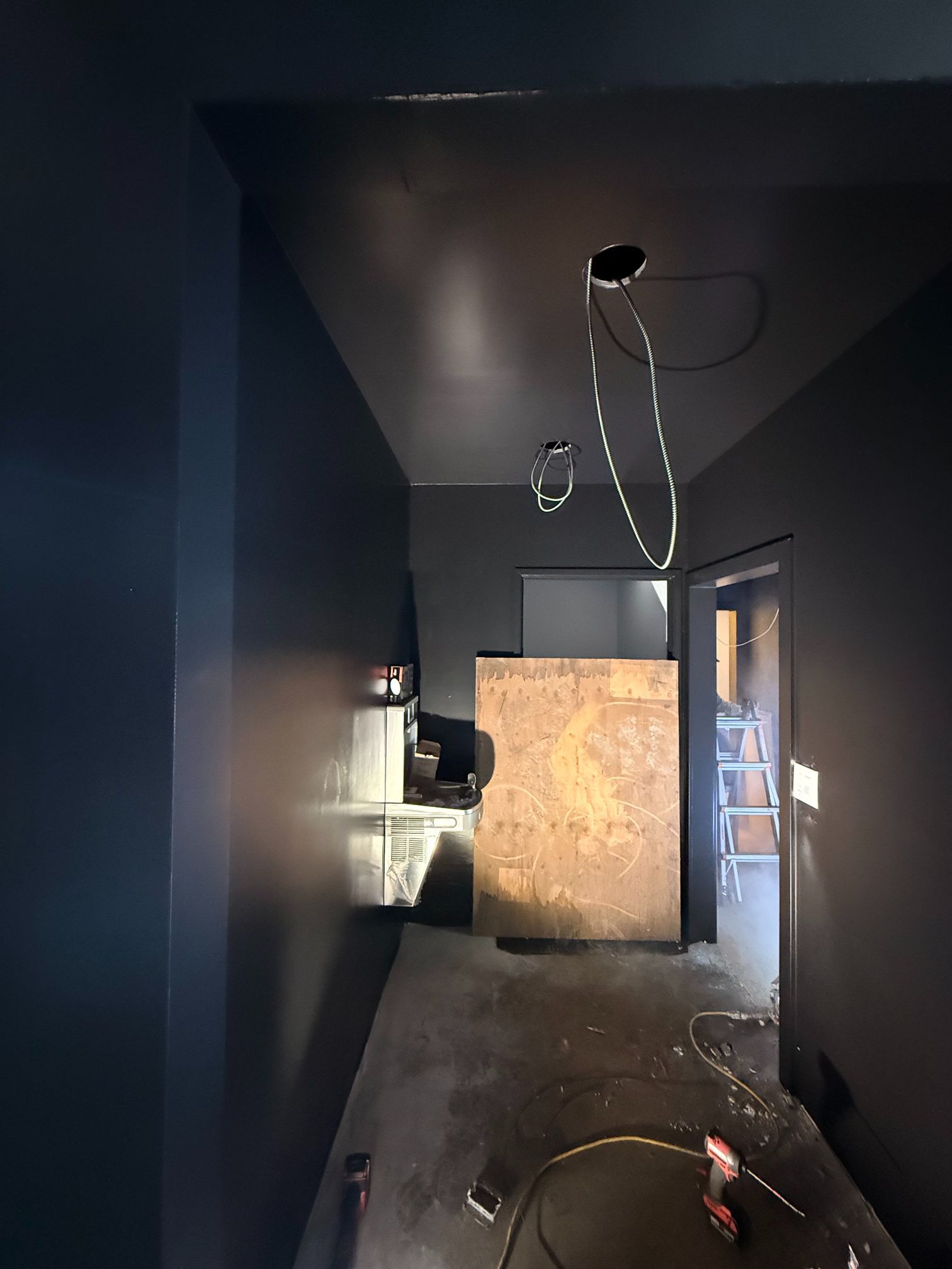 Dim unfinished hallway with dark walls, exposed wiring, and a plywood board blocking the far end.
