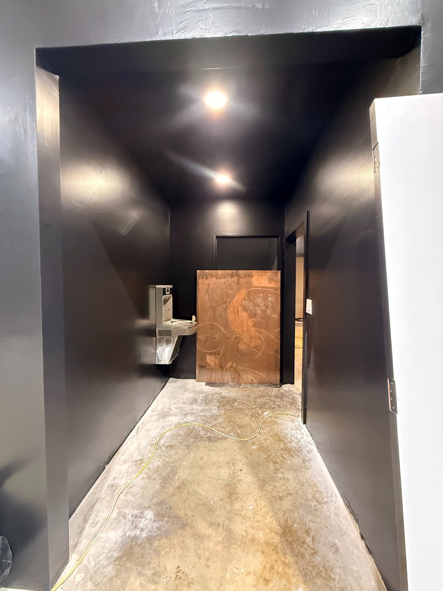 Narrow dark hallway with black walls, concrete floor, and a rusty door at the end.