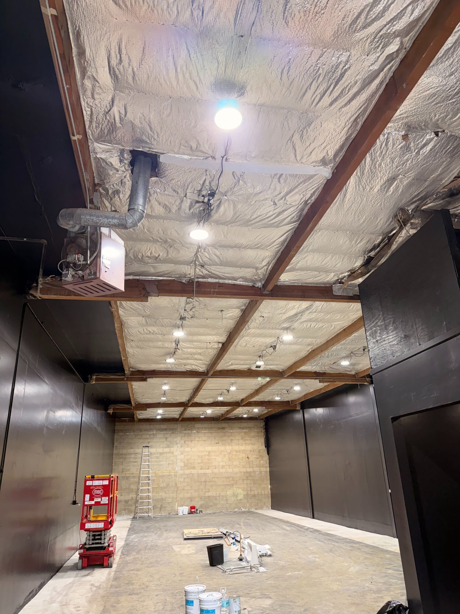 Empty industrial room with foil-insulated ceiling, black walls, exposed beams, and construction materials on the floor