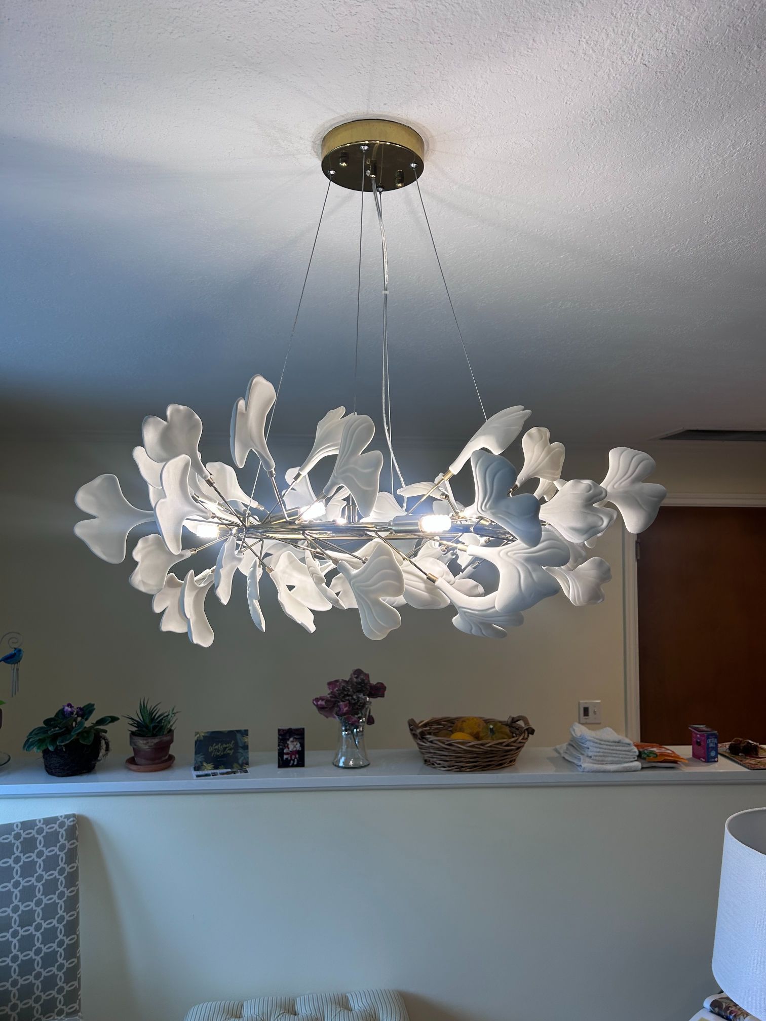 White floral chandelier hanging from a ceiling in a room, with shelf decor and a lamp below.