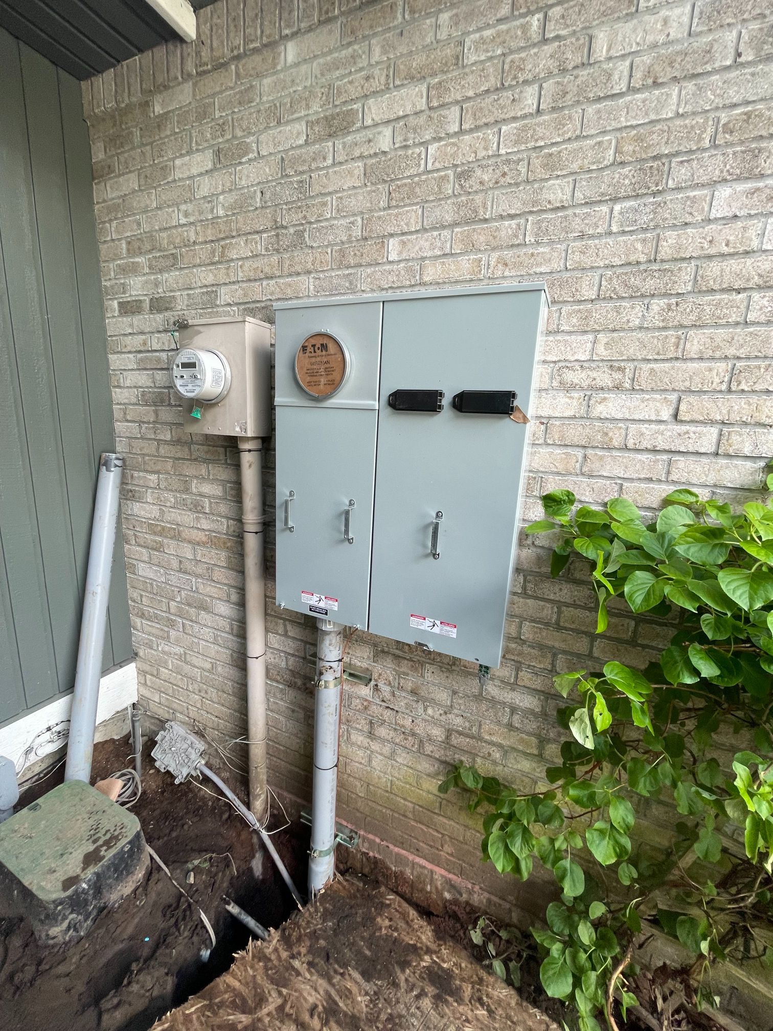 Two gray utility boxes on a brick wall with pipes, an electric meter, and exposed trenching below.