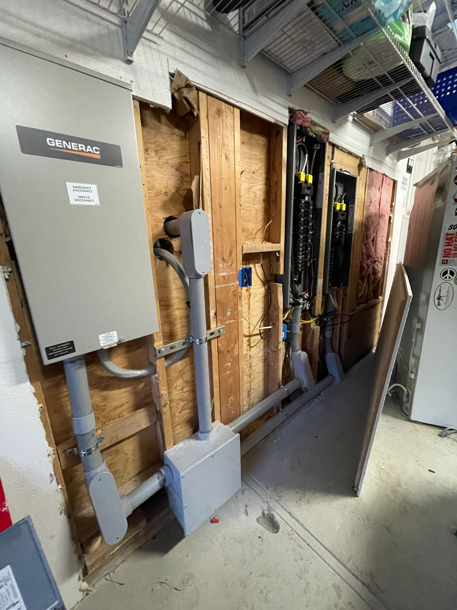 Damaged utility wall with exposed framing, pipes, electrical panels, and a leaning panel in a narrow corridor.