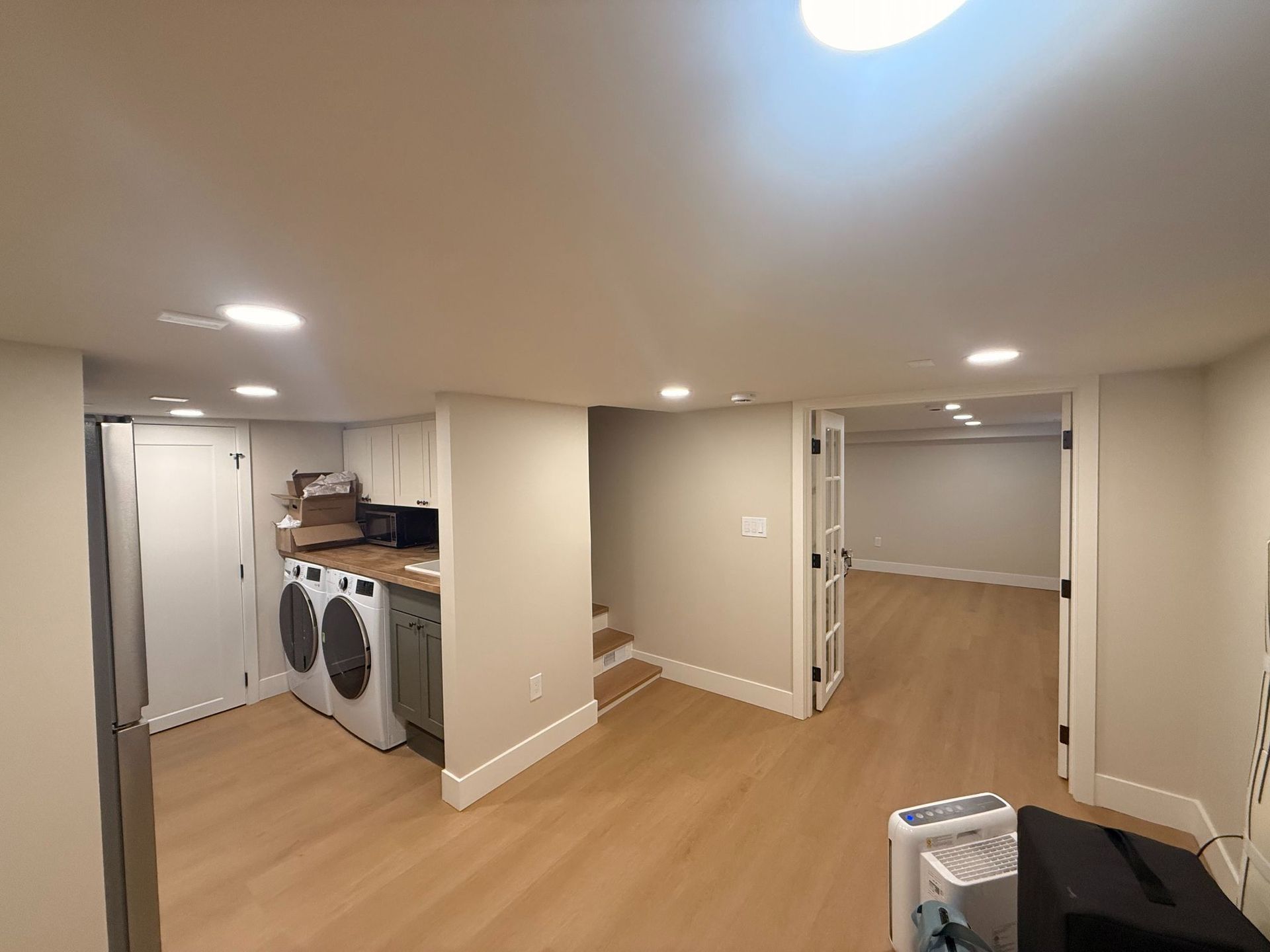 Empty basement laundry room with wood floors, recessed lights, and adjacent open storage area.
