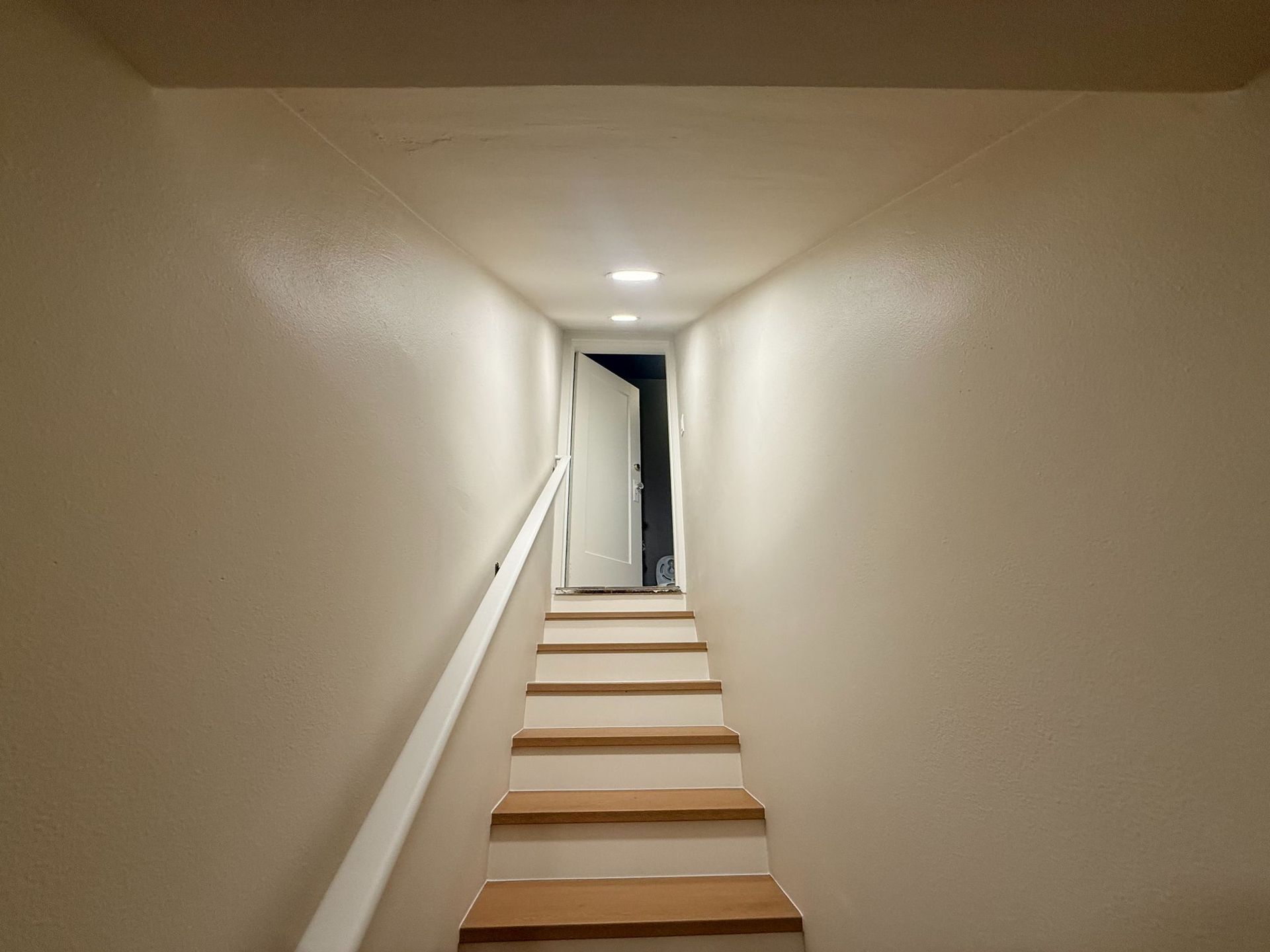 Narrow stairwell with beige walls and white steps leading up to an open doorway.