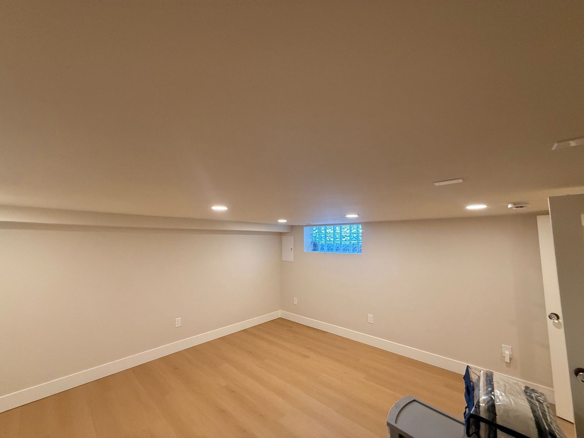 Empty beige basement room with wood floor, recessed lights, and a small window.