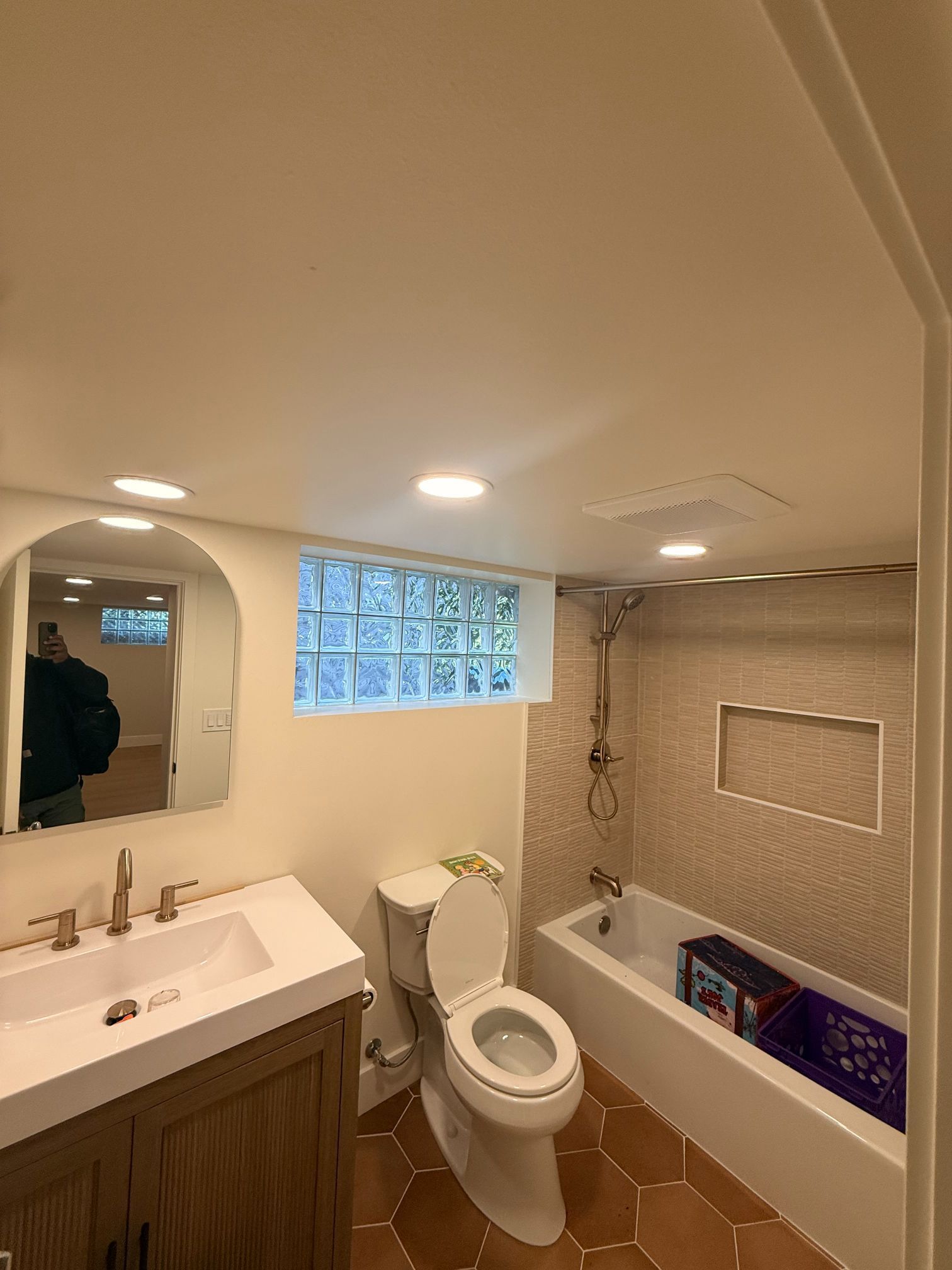 Small beige bathroom with sink, toilet, tub, mirror, and frosted window; a person is reflected in the mirror.