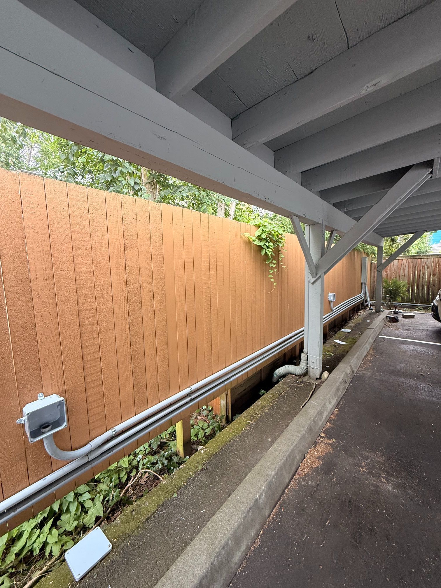 Covered walkway beside a tan fence and drainage channel, with gray beams and wet pavement.