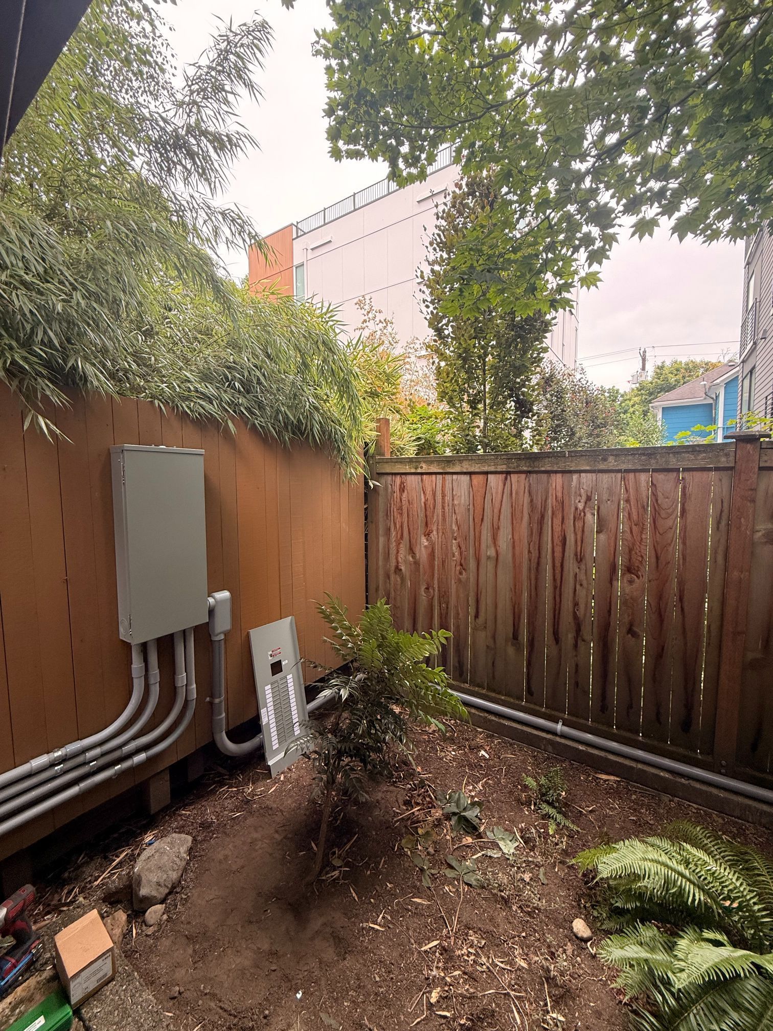 Backyard utility area with electrical boxes on a wooden fence, dirt trench, and surrounding greenery