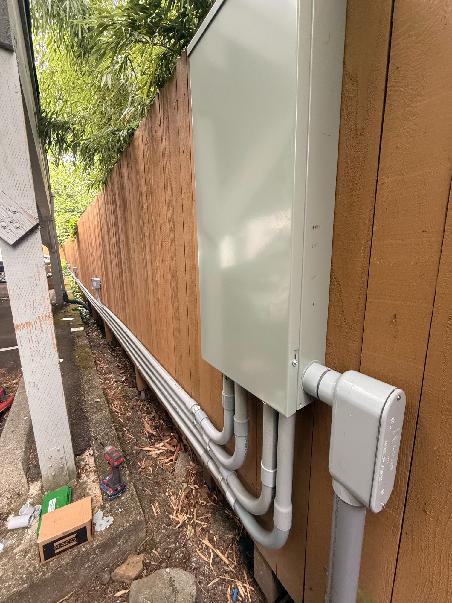 Outdoor utility conduits and a white electrical box mounted along a fence beside a narrow walkway