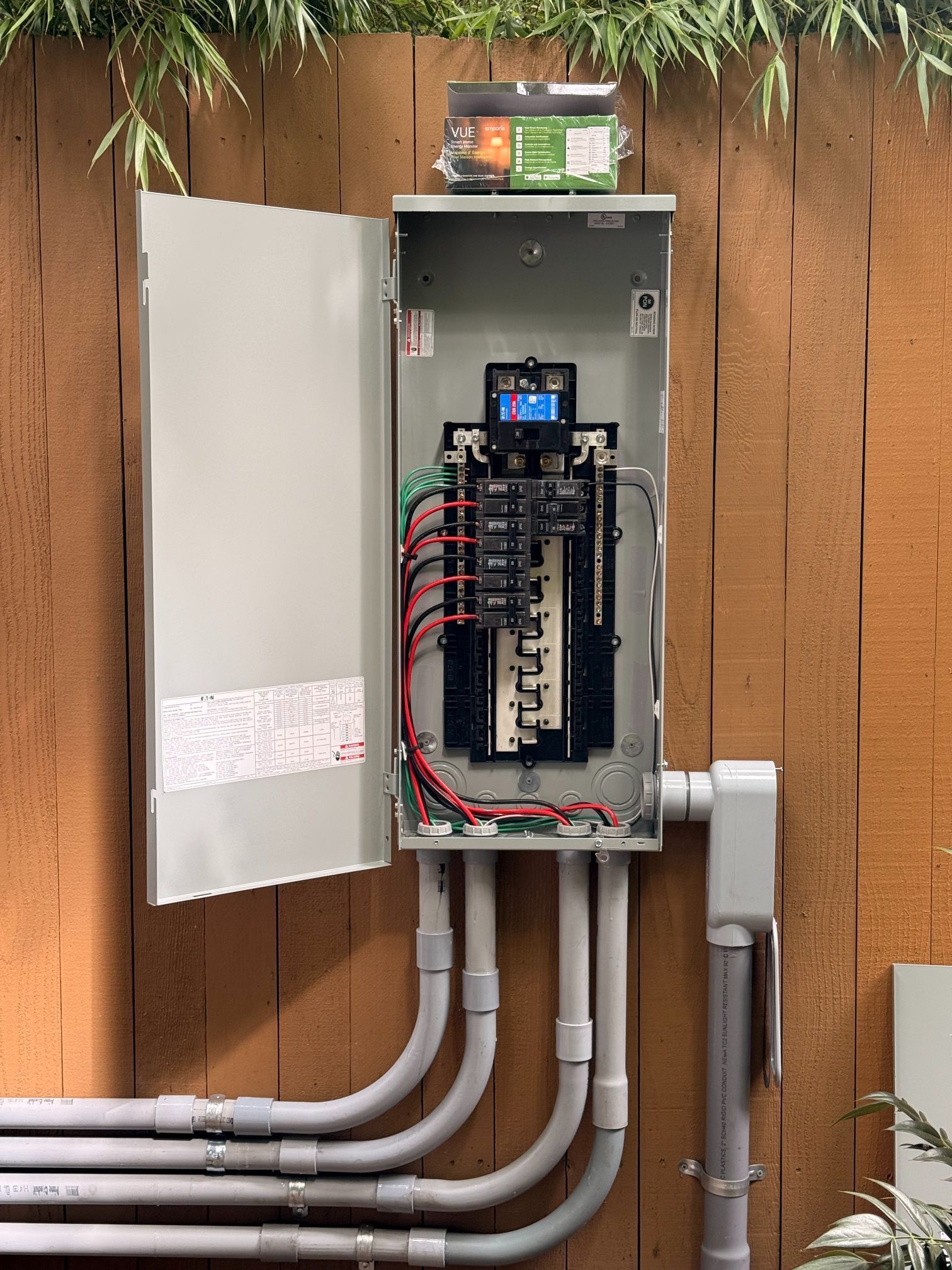 Open electrical panel on exterior wall with exposed wiring and conduit, near wooden fence and plants