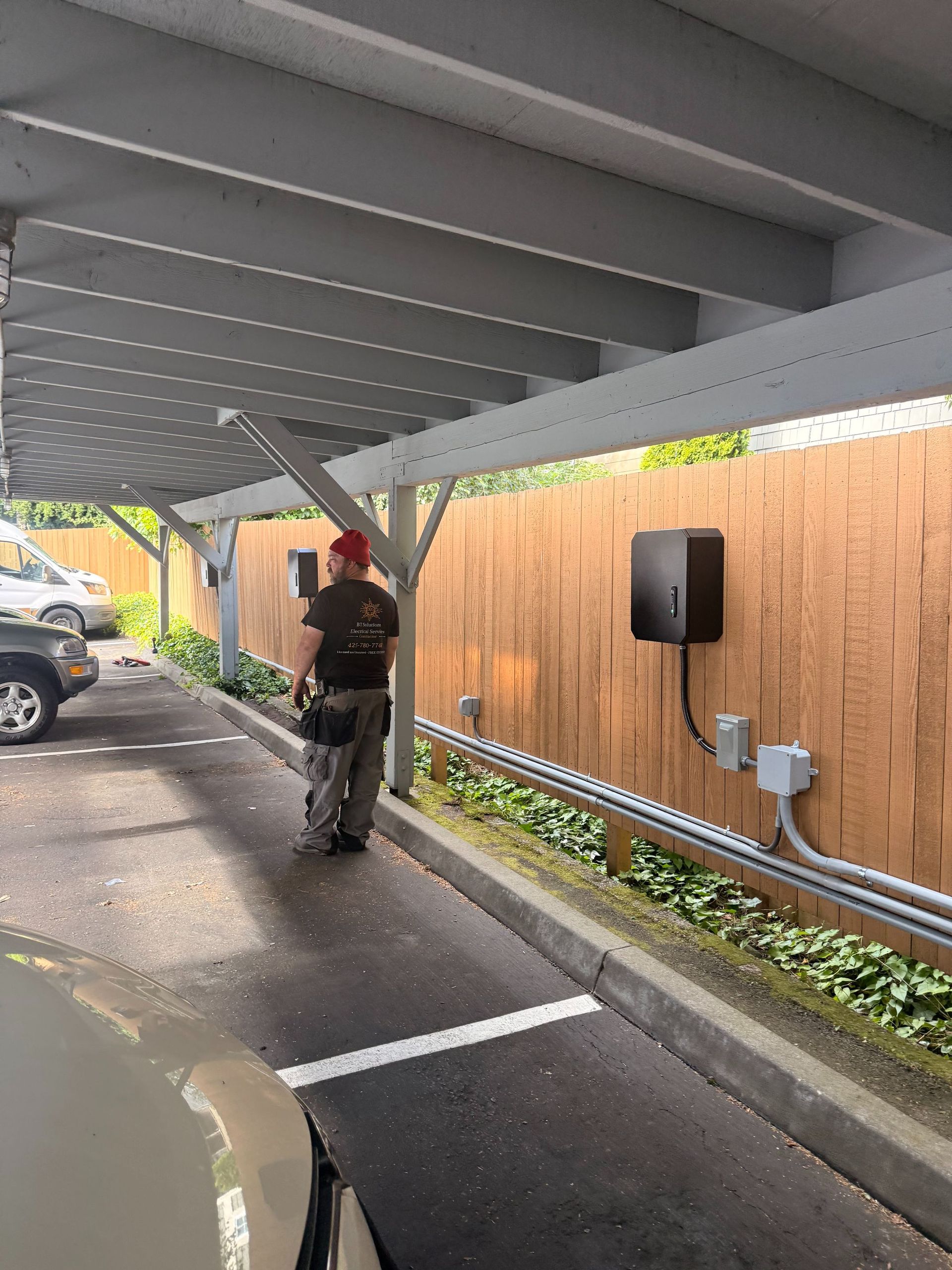 Worker walking under a covered parking area beside a wooden fence and parked vehicles.