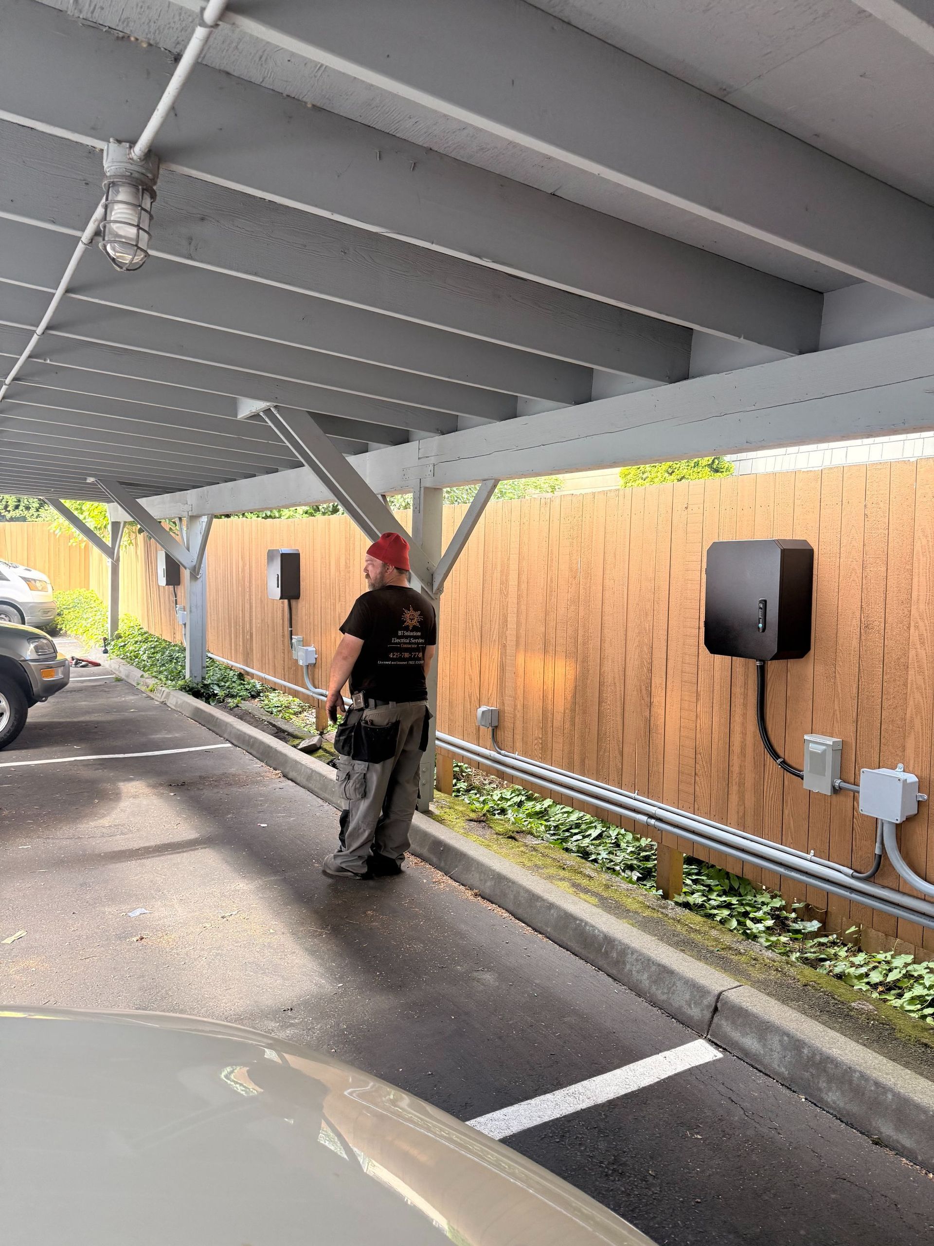 Worker standing under a covered parking area next to a wall-mounted charging box, holding equipment.