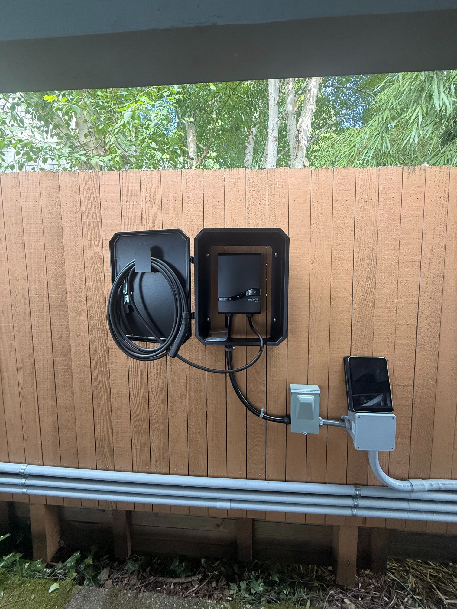 Outdoor wall-mounted electrical boxes and coiled cable on a wooden fence, with conduit below.