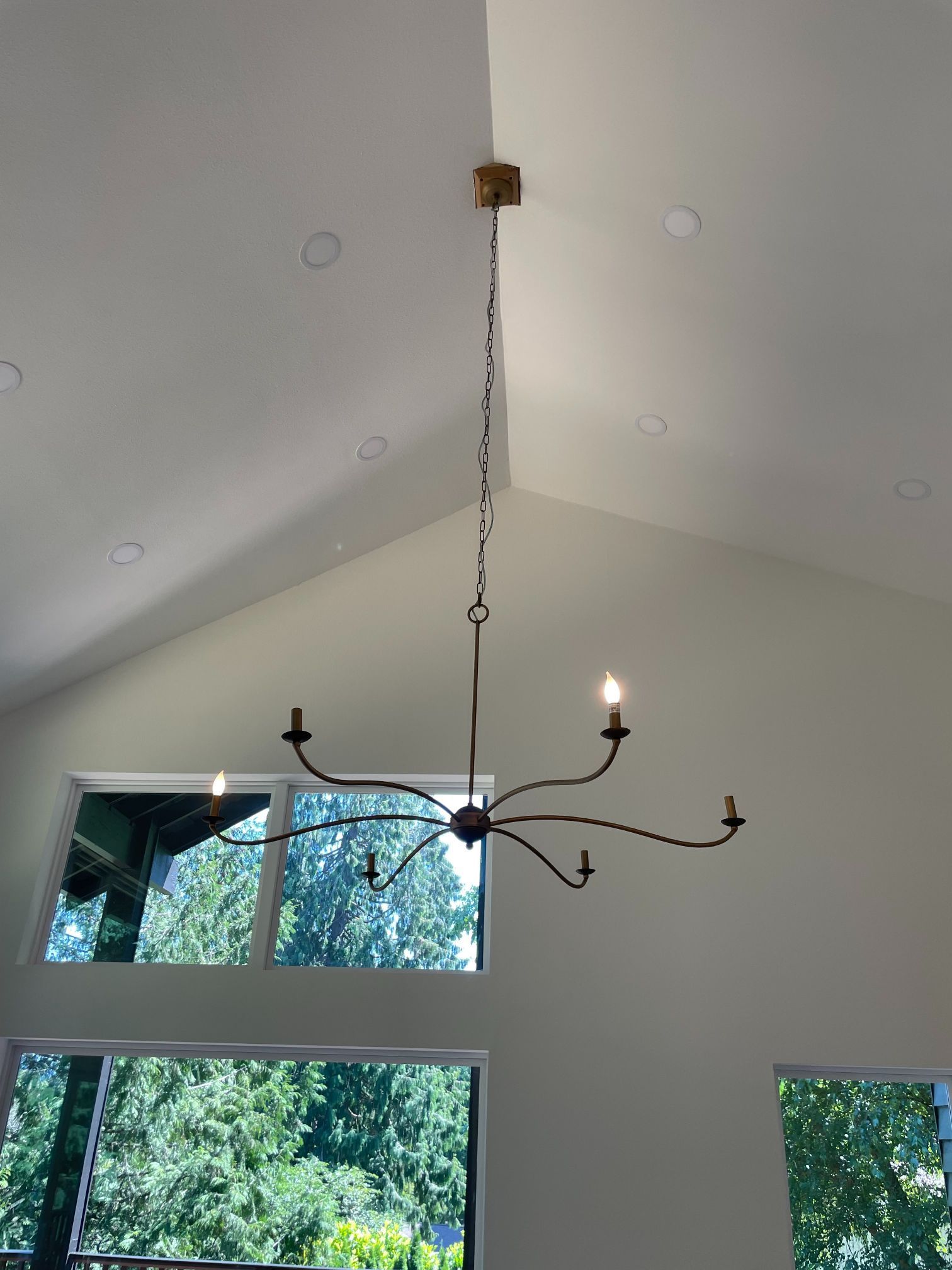 Chandelier hanging from a high white ceiling above large windows with trees outside.