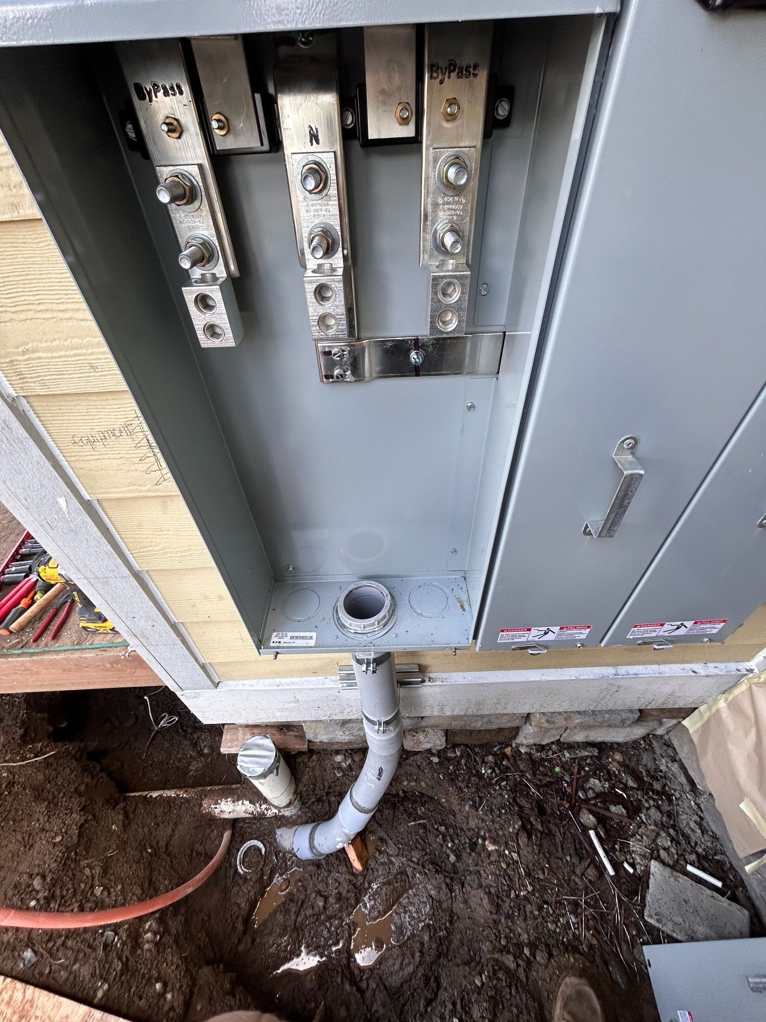 Open electrical panel with exposed wiring and conduit above a dirt trench outdoors