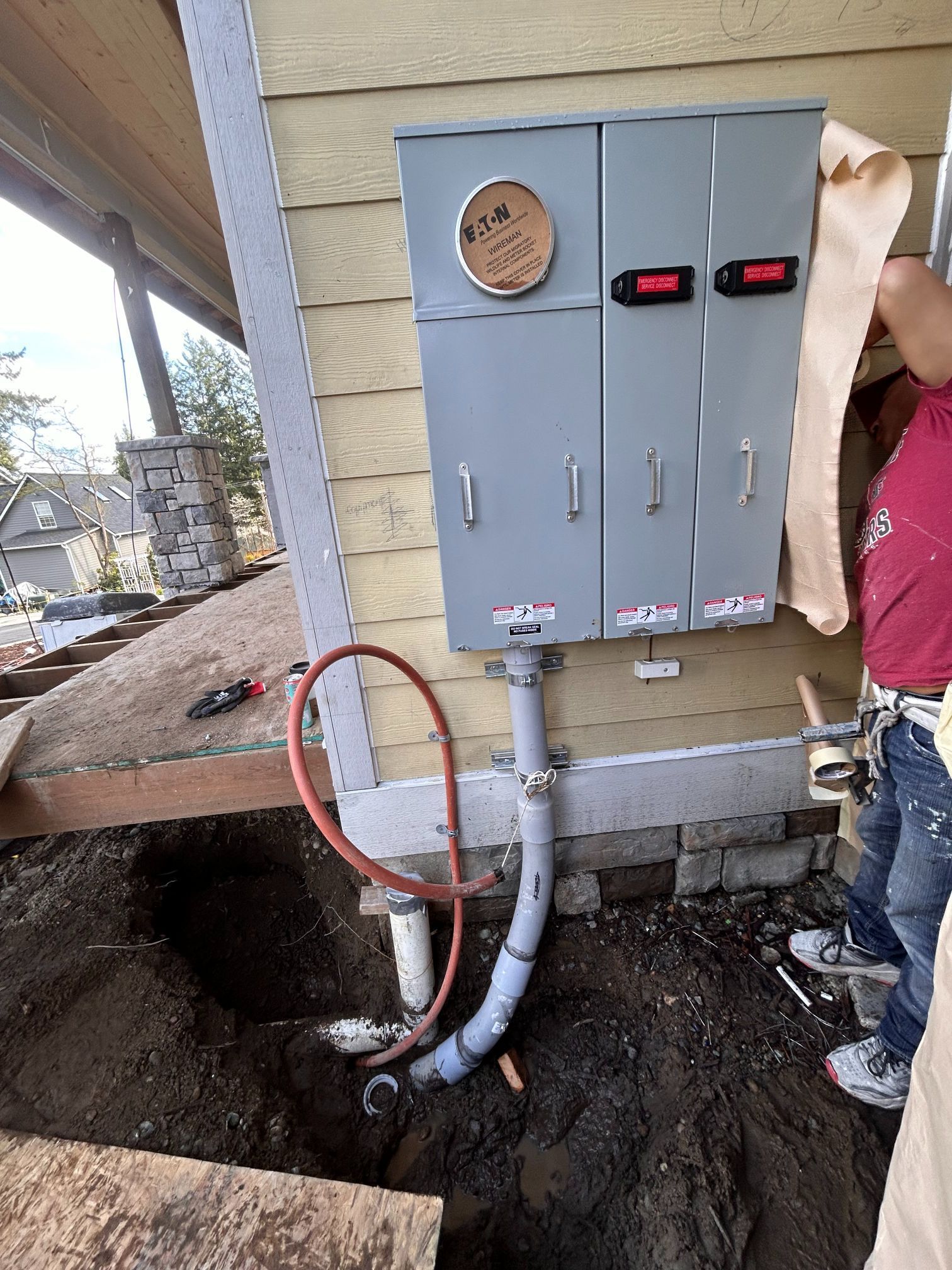 Outdoor electrical meter and gray service panel mounted on a house beside an open trench and exposed cables.