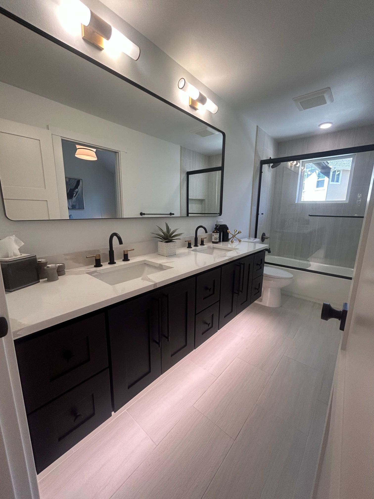 Modern bathroom with double vanity, large mirror, black cabinets, and white countertops; window at the far end