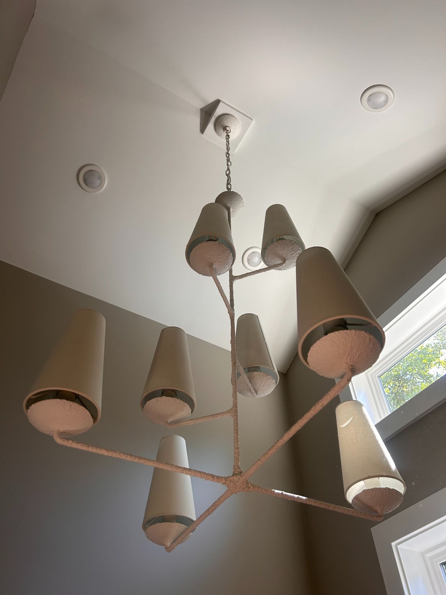 Ceiling light fixture with several beige lampshades on a white ceiling near a window.