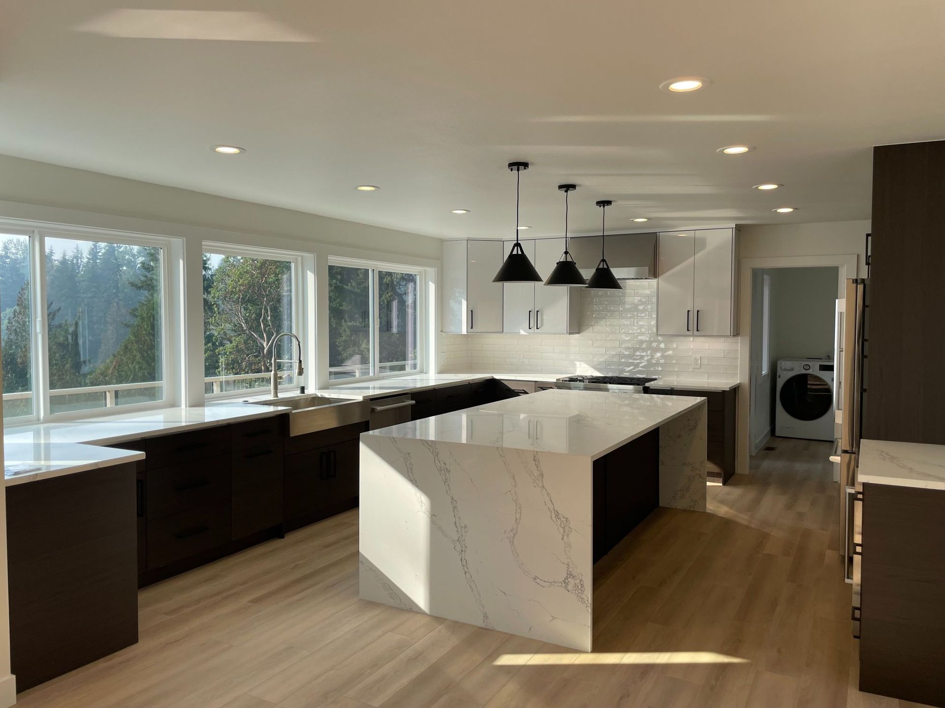 Modern kitchen with white marble island, dark cabinets, pendant lights, and large windows overlooking trees