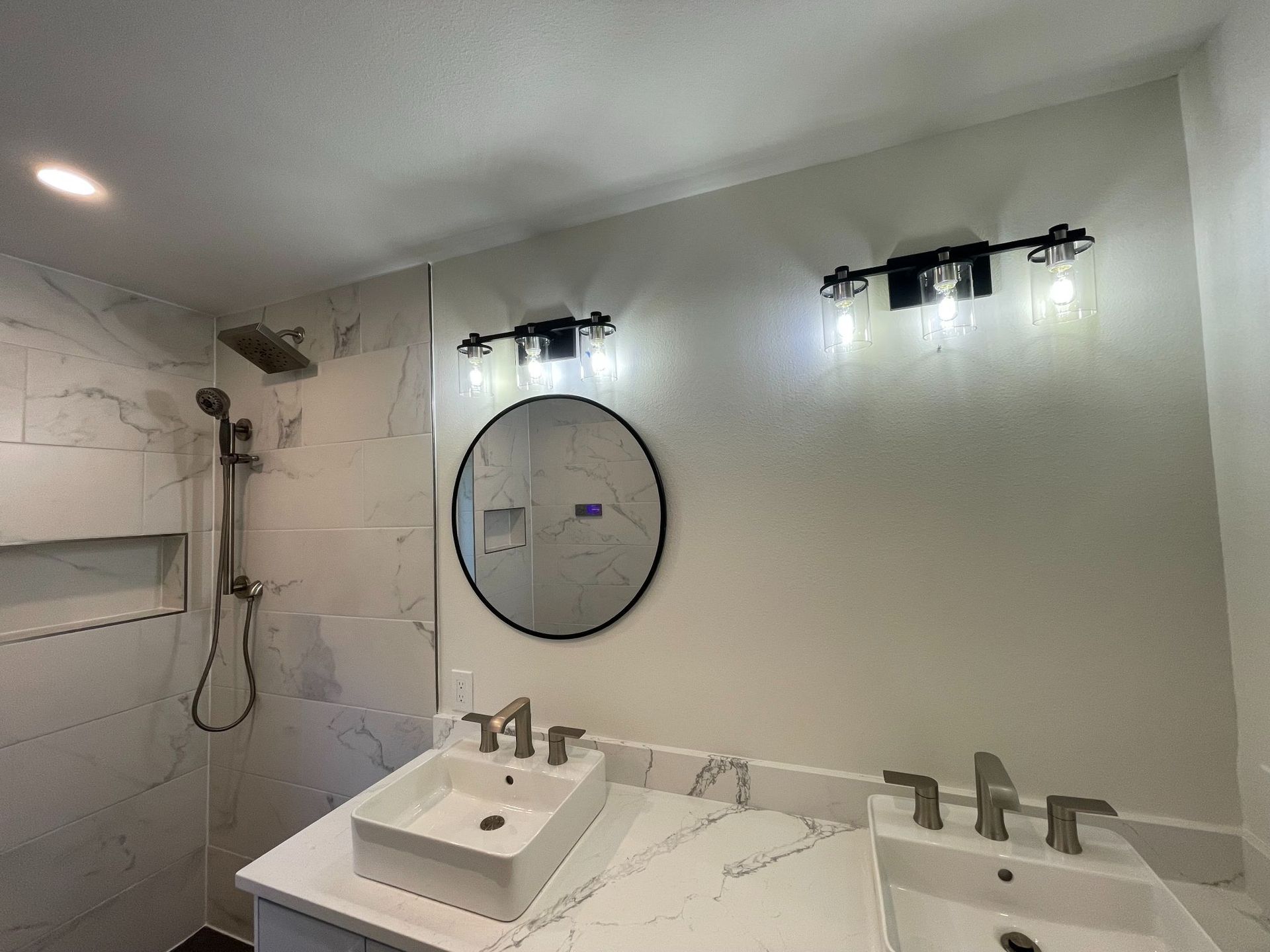 Modern bathroom with double sink vanity, round mirror, wall lights, and shower stall