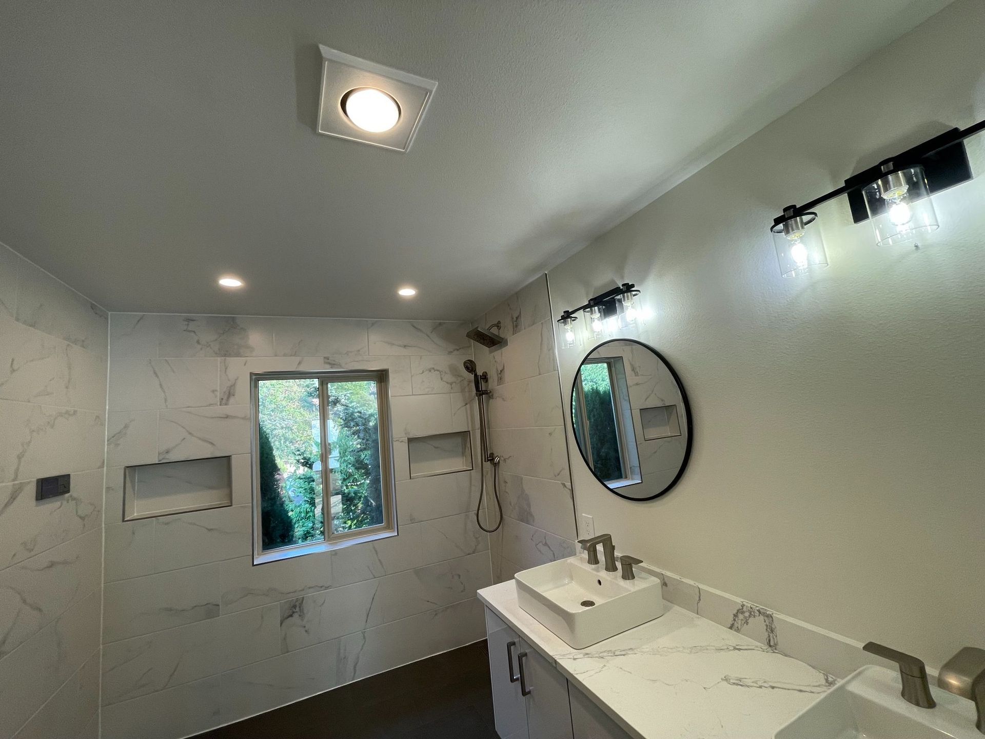 Modern bathroom with white marble walls, double sink vanity, round mirror, and window above the bathtub area