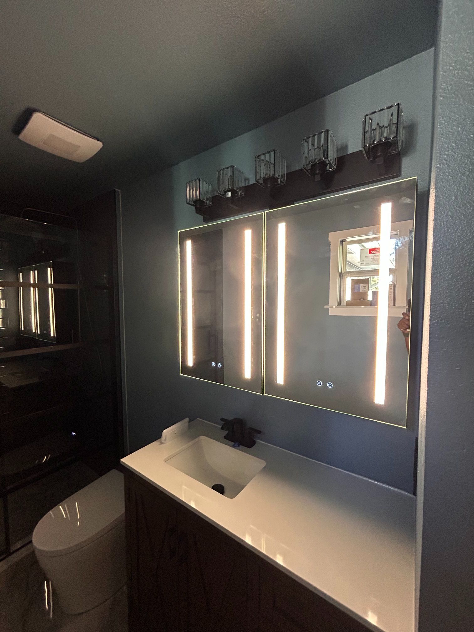 Modern bathroom with lit mirror, white sink, dark walls, and toilet in the foreground