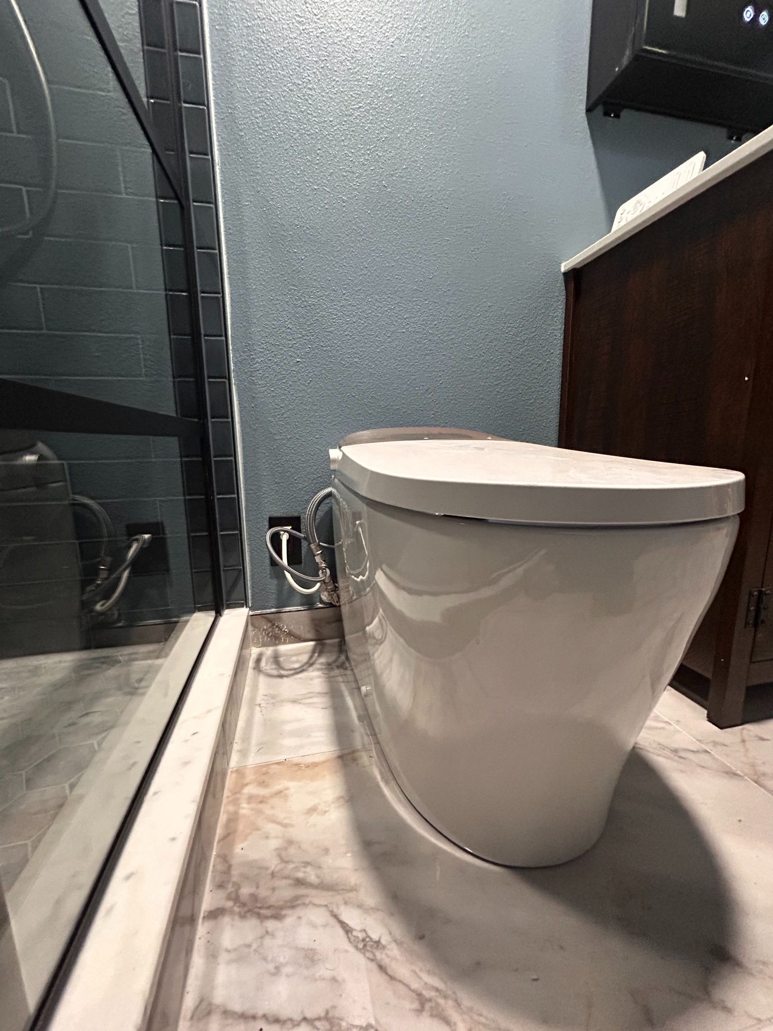 Modern bathroom with a white toilet beside a glass shower and dark vanity on marble tile.