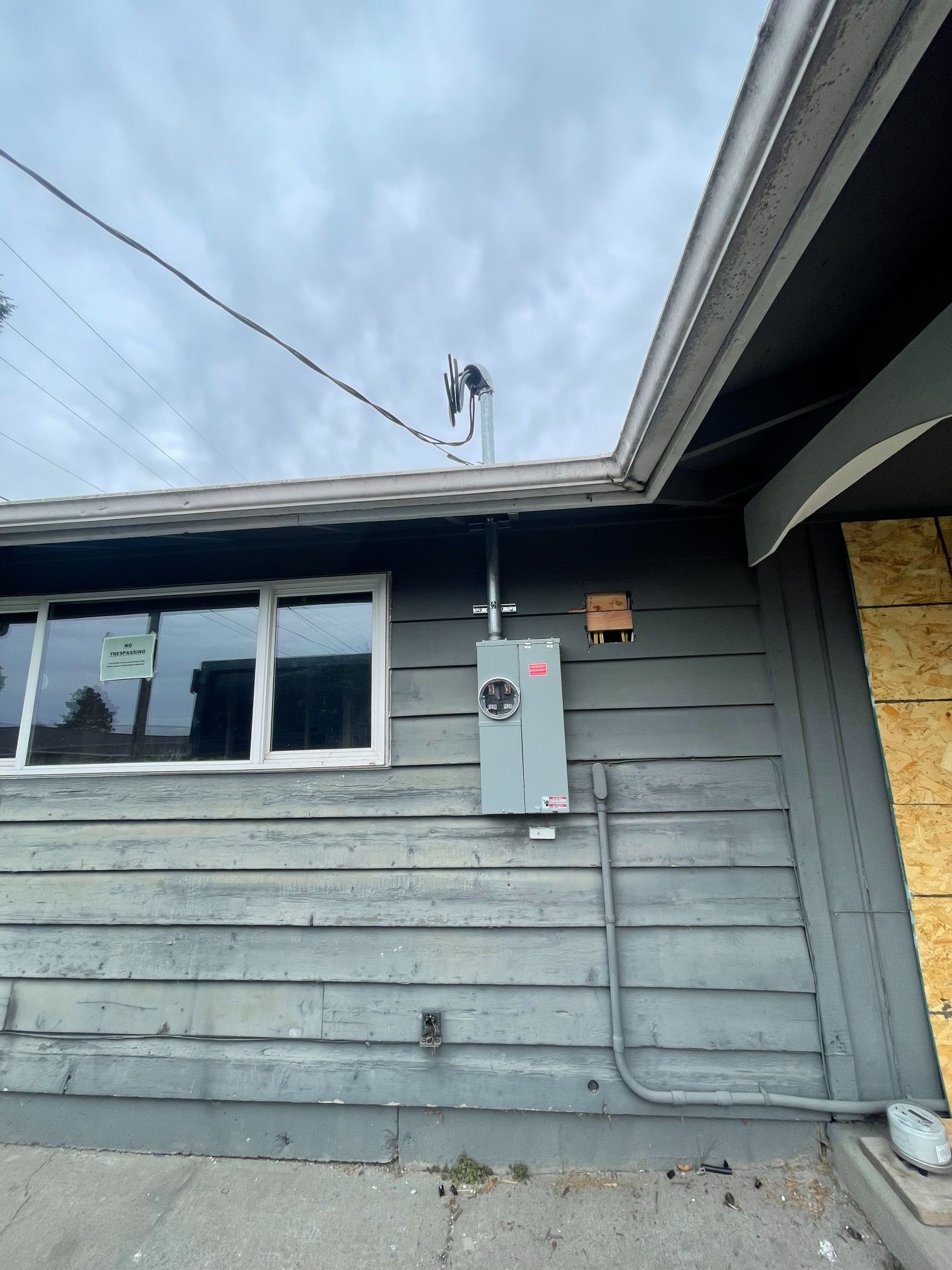 Exterior of a gray house wall with window, electrical panel, and roofline under cloudy sky