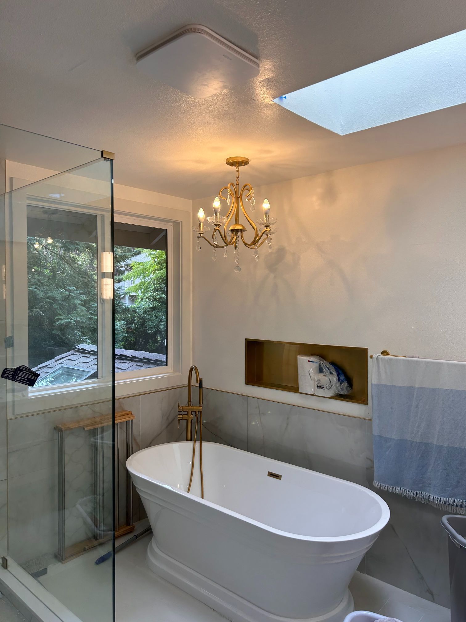 Modern bathroom with freestanding tub, glass shower, chandelier, skylight, and window view