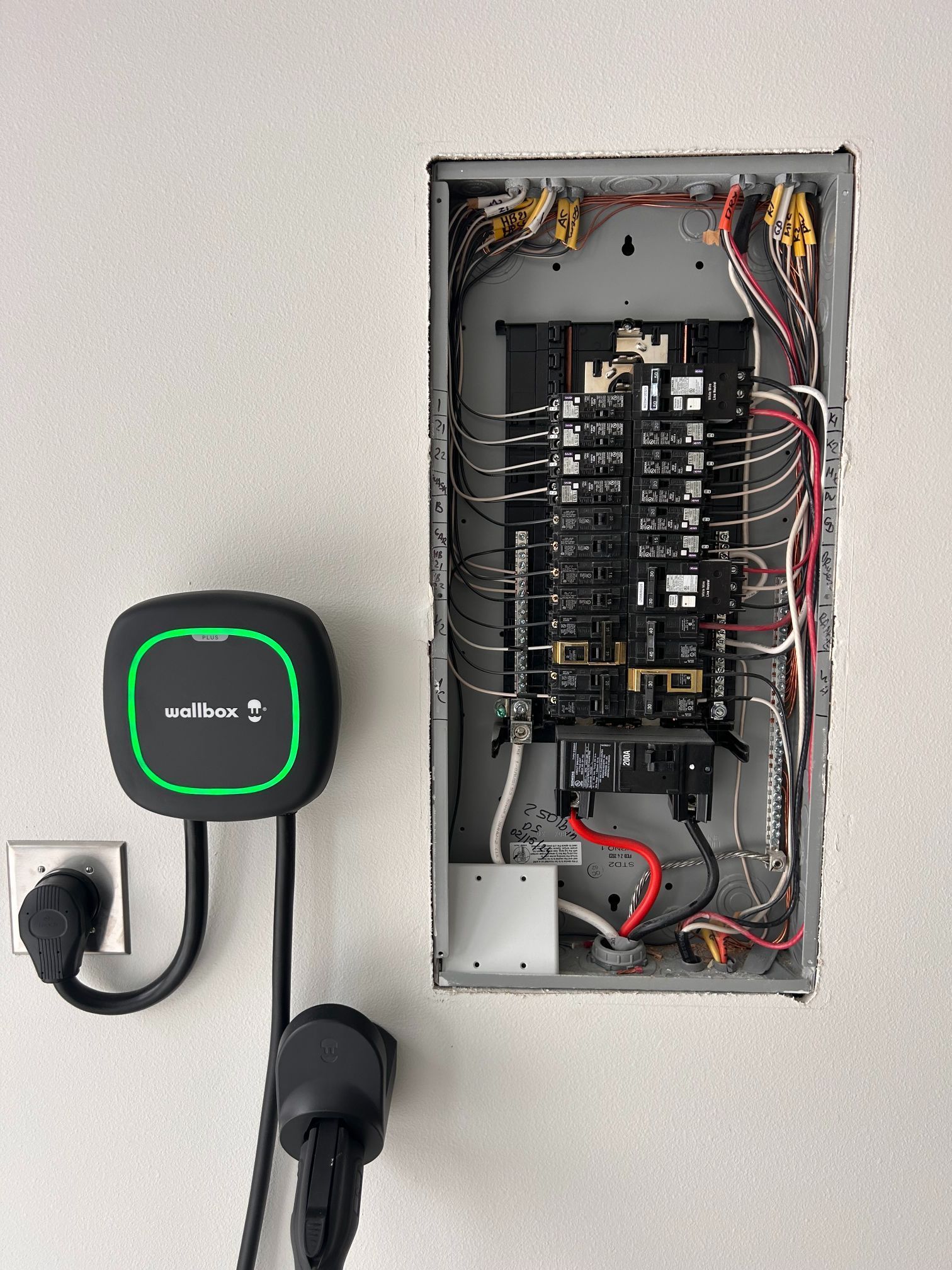 Open electrical breaker panel next to a wall-mounted black charger with green indicator light.