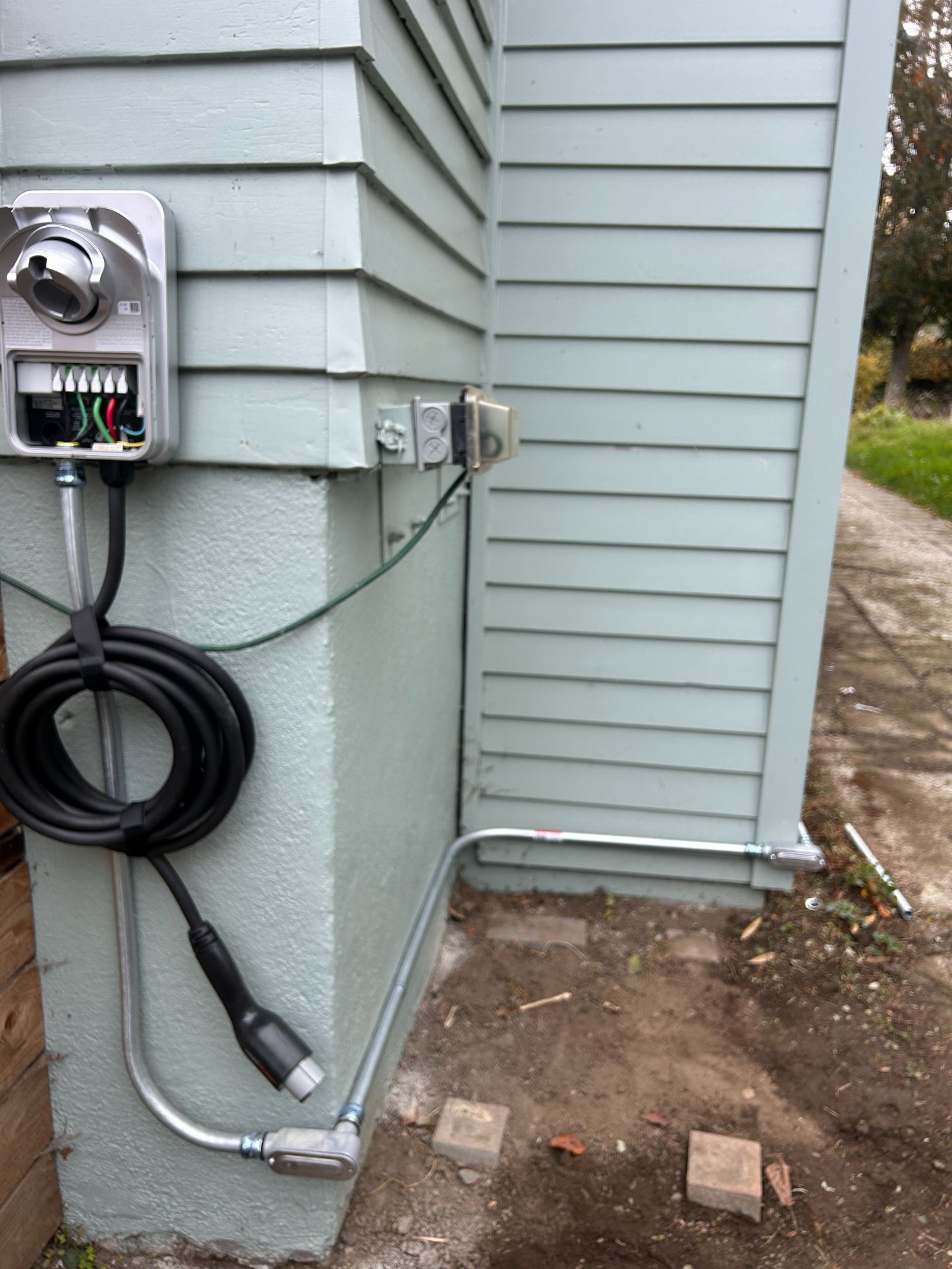 Outdoor electrical meter and charging cable mounted on a house exterior near a dirt path.